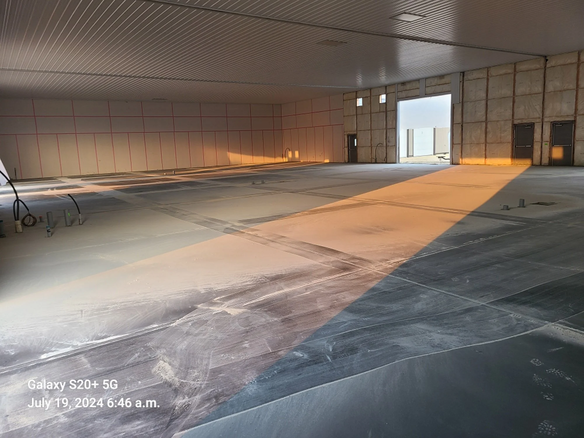 An interior of a building under construction with exposed insulation walls, a concrete floor, and large open doorway with sunlight streaming in.