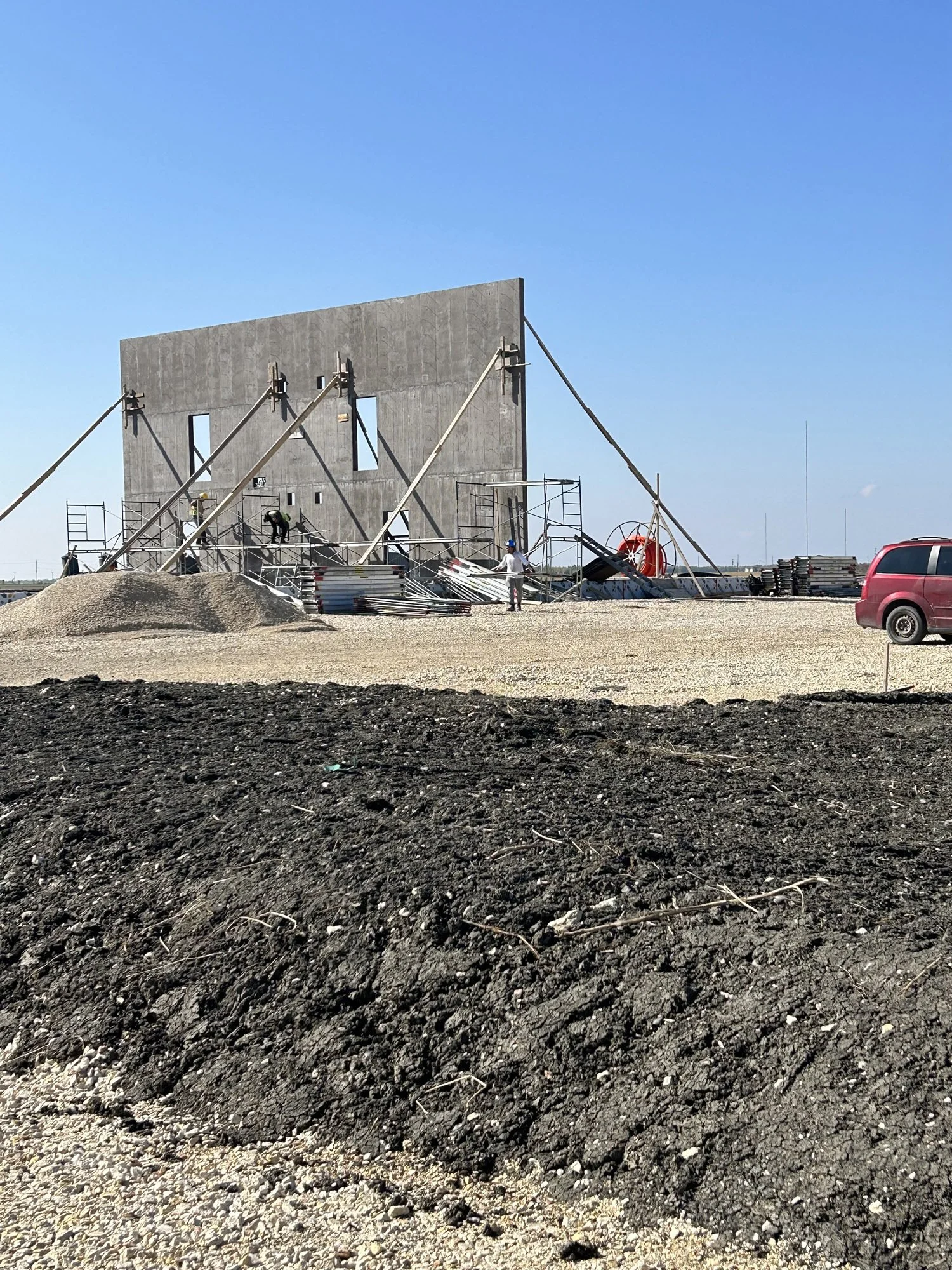 Construction site with workers building a large gray concrete wall, supported by diagonal beams, with a clear blue sky in the background. There are construction tools and materials, including scaffolding, gravel, and debris, along with a parked red m
