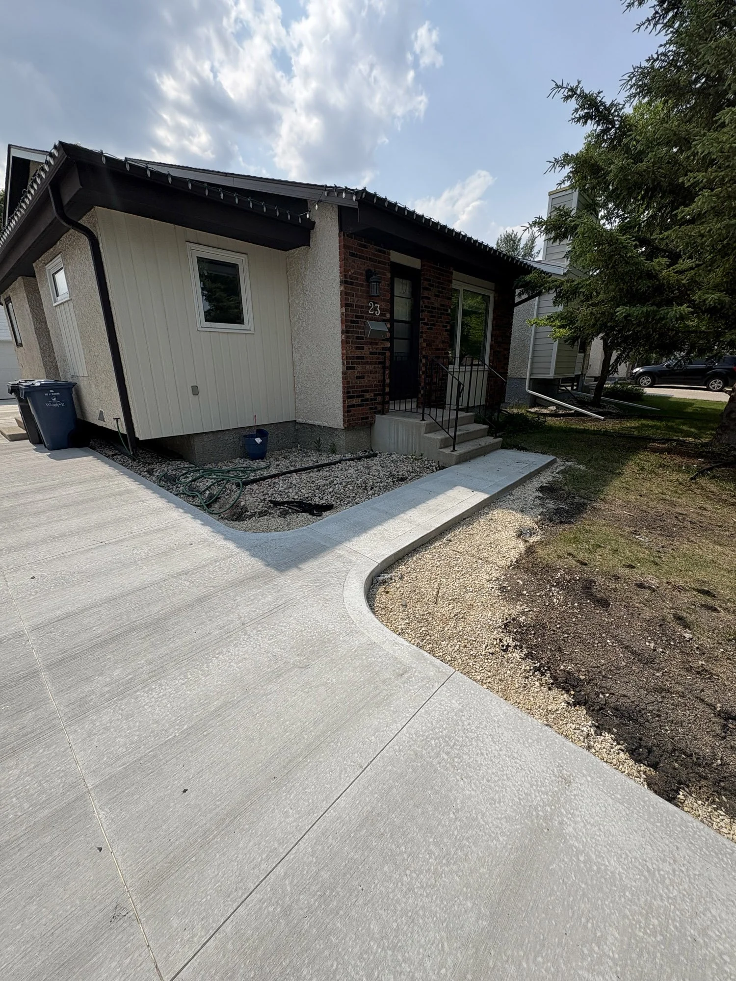 Front view of a modern house with a concrete driveway, steps leading to the front door, and landscaping work in progress, including gravel and dirt areas, trees, and neighboring houses in the background.