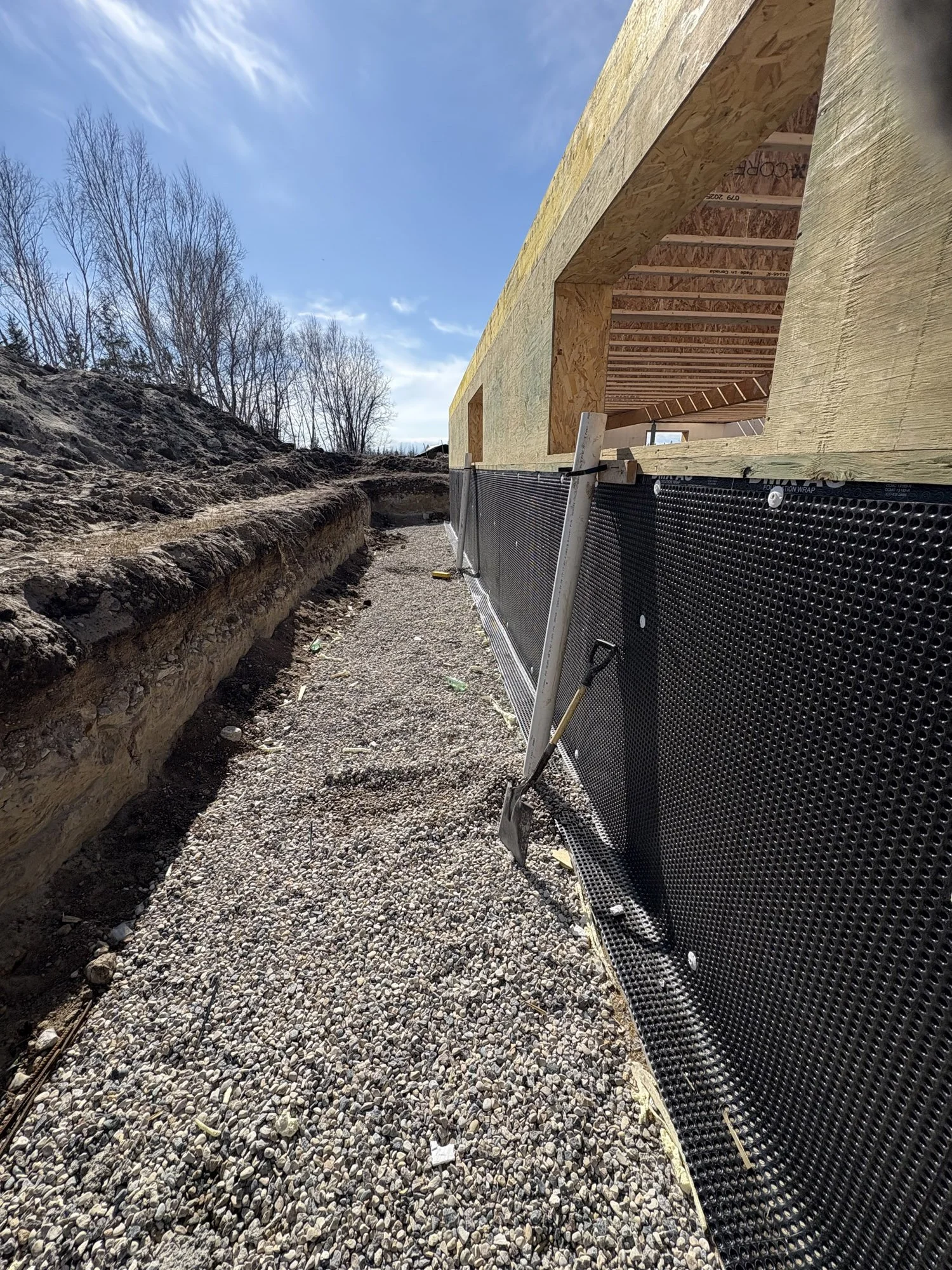 Side view of a house under construction with plywood walls, black foundation wrap, and gravel ground, next to a dirt excavation site with leafless trees and a blue sky.