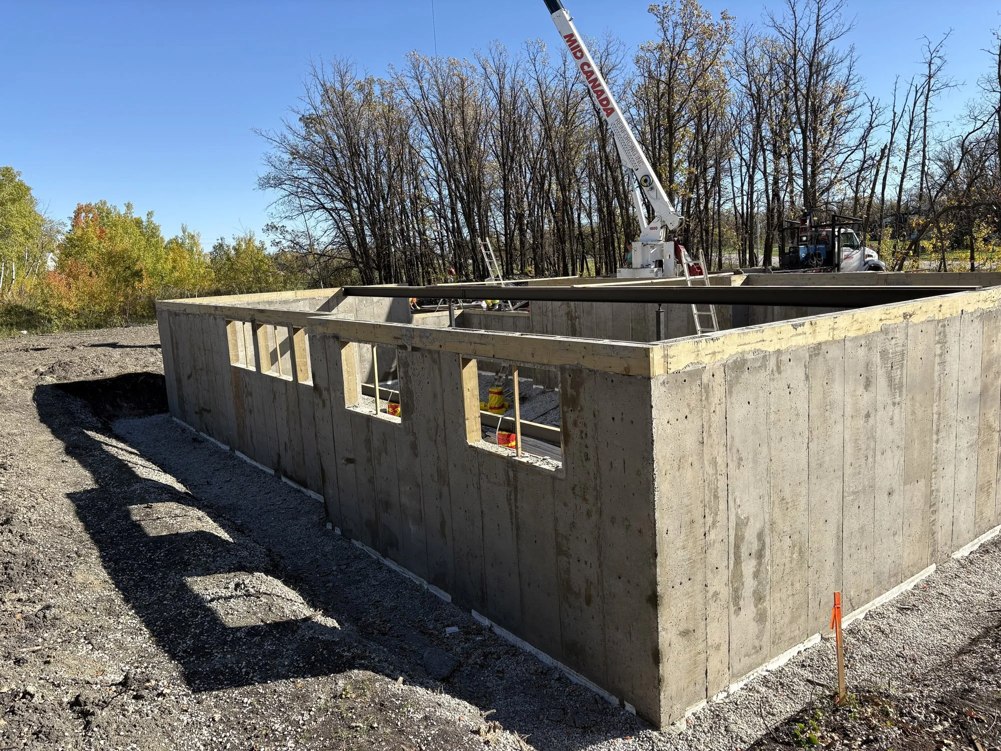 Construction site with a concrete foundation and steel rebar framework, a crane, and construction vehicles, set against trees with fall foliage on a sunny day.