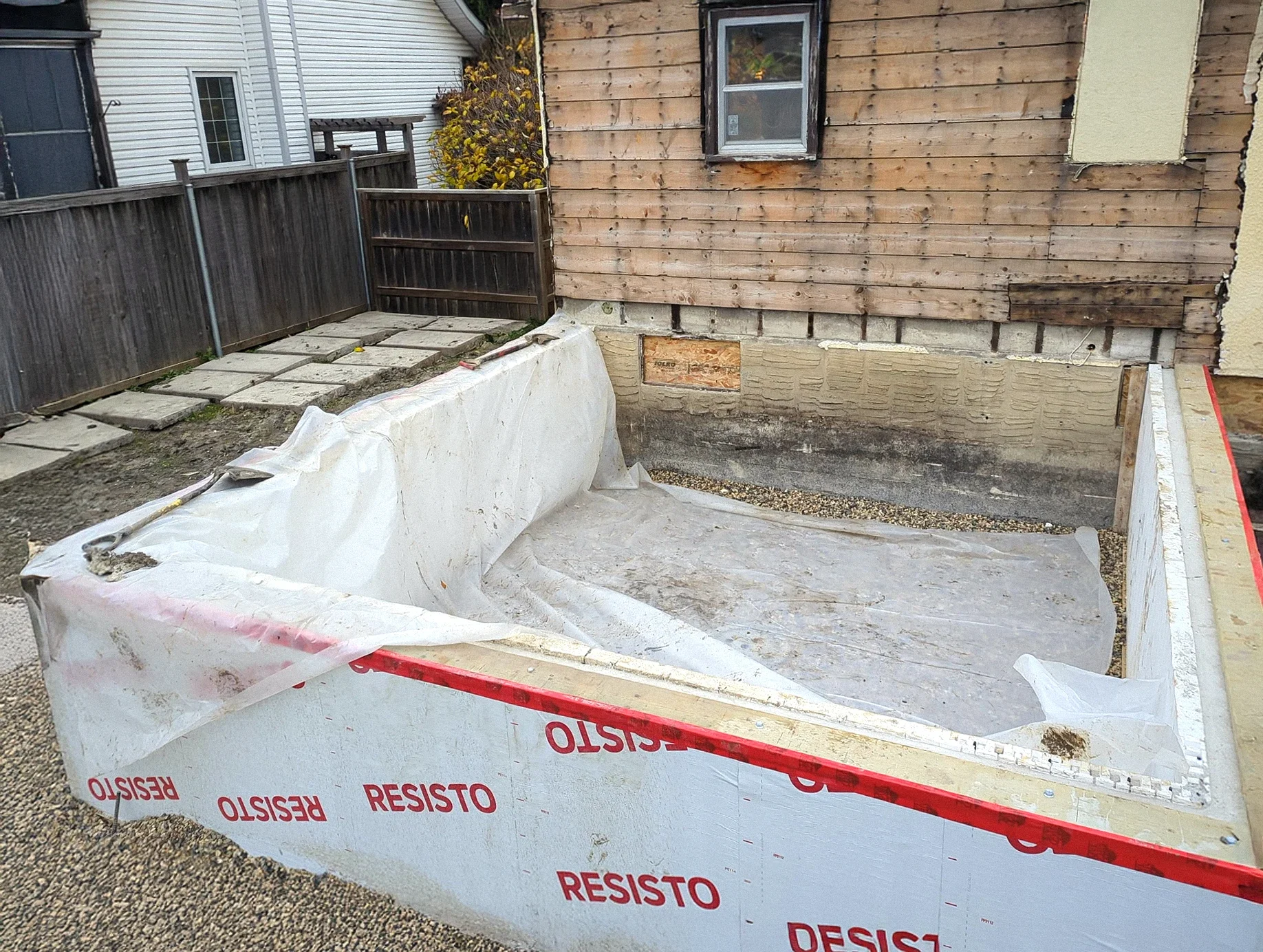 Backyard construction site, surrounded by a white form with red RESISTO branding, wooden fencing, and a house with wooden siding and a small window.