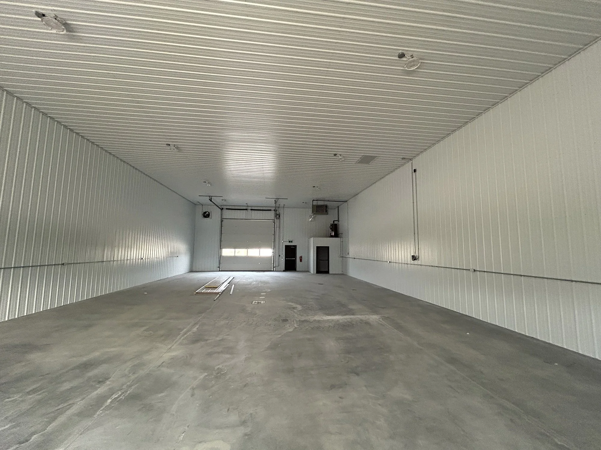 Empty industrial warehouse with white metal walls and ceiling, concrete floor, and a large garage door at the back.