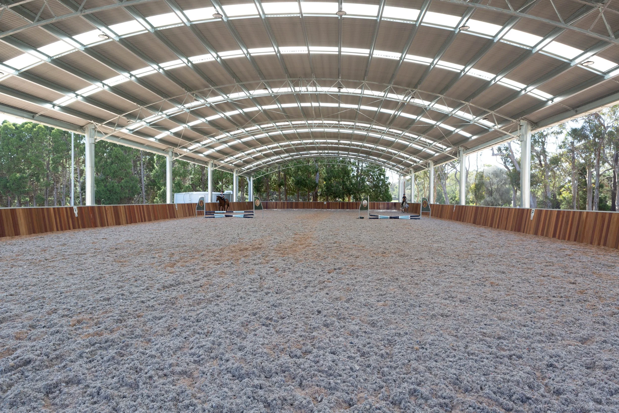 An indoor equestrian arena with a sand riding surface, covered by a curved metal roof supported by white pillars, surrounded by a wooden barrier, with trees visible outside.