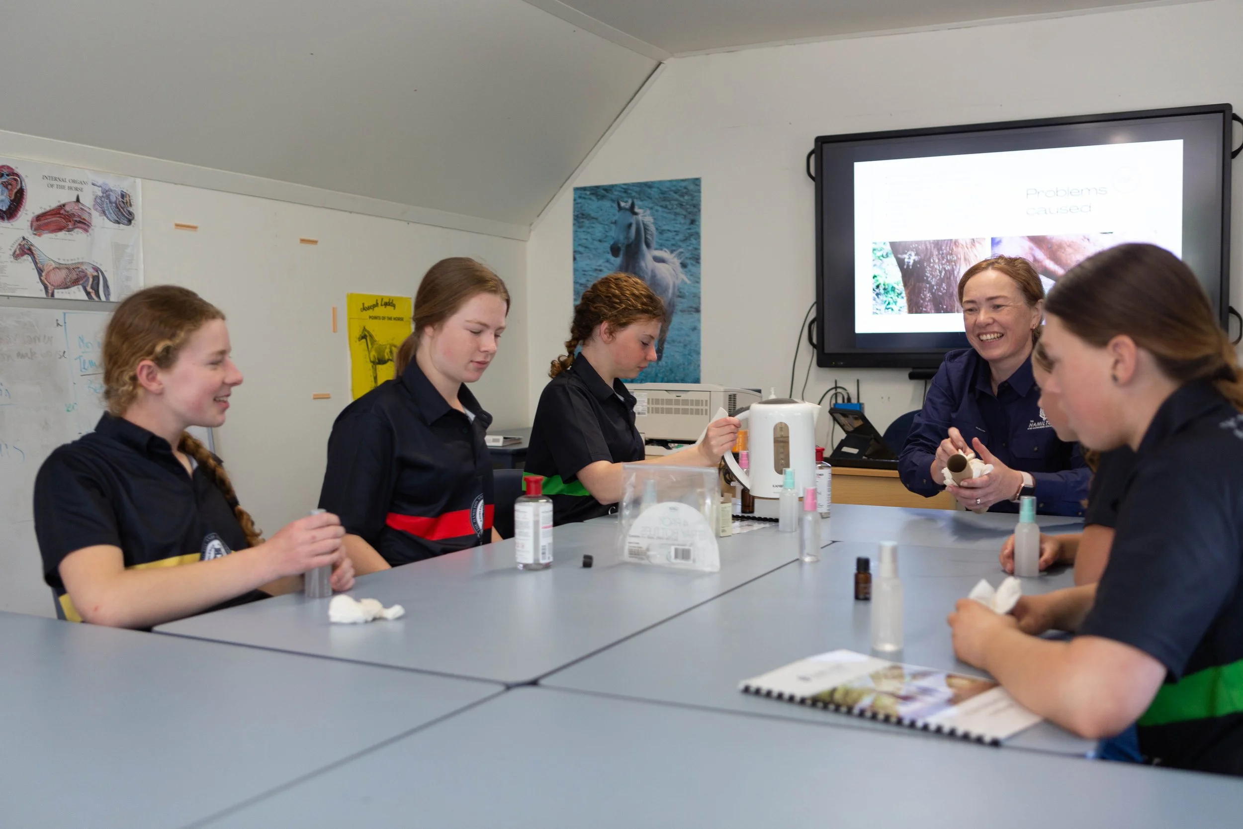 A group of five women sitting around a table in a classroom or workshop, engaging in a nail polish or beauty activity. They are smiling and focused on their tasks, with nail polish bottles and tissue paper on the table. There is a large screen on the