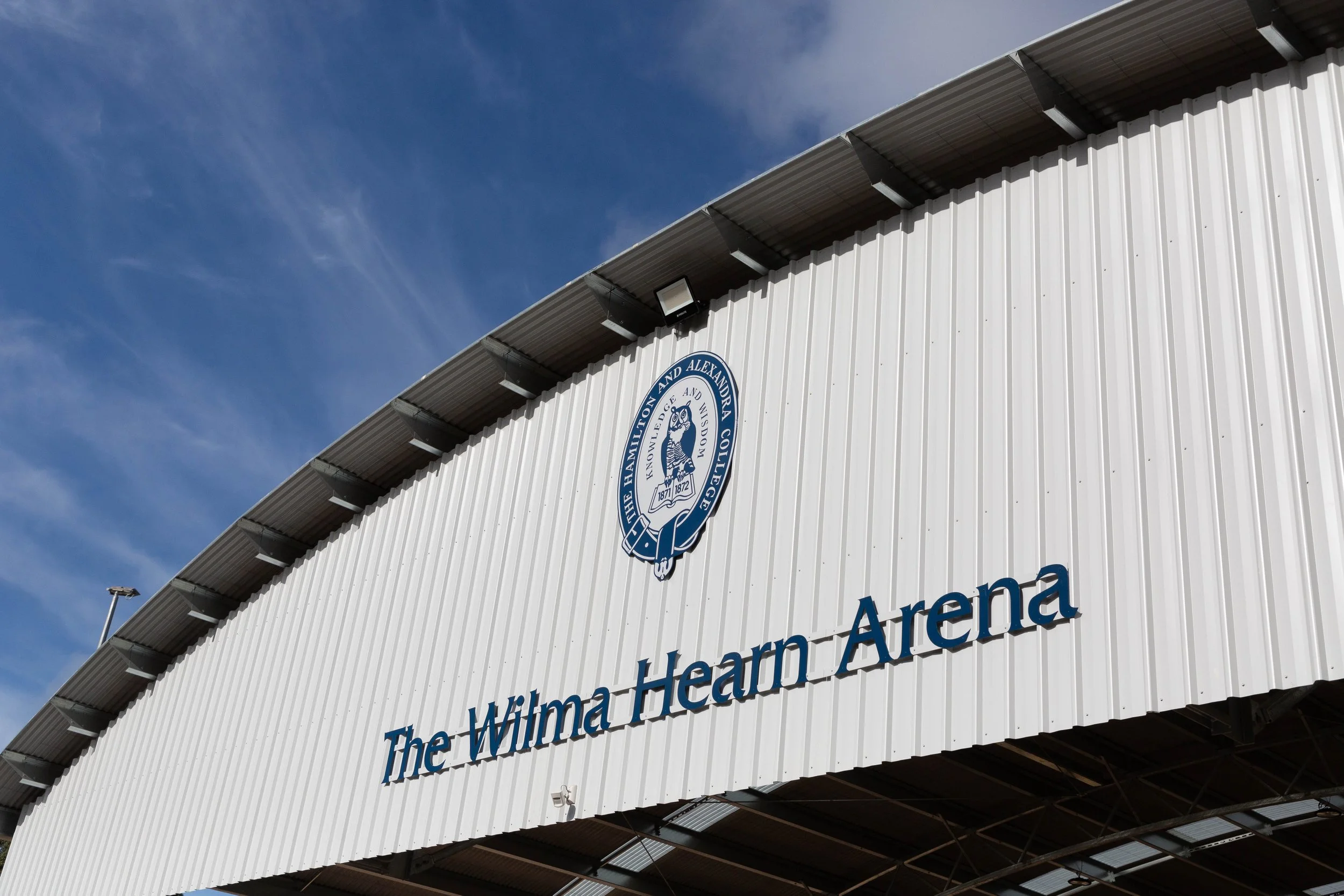 Part of the exterior of a sports arena with a large sign that says 'The Wilma Wima Hearn Arena' and a school crest logo above it. The building has a white corrugated metal facade and a sloped metal roof, with clear blue sky in the background.