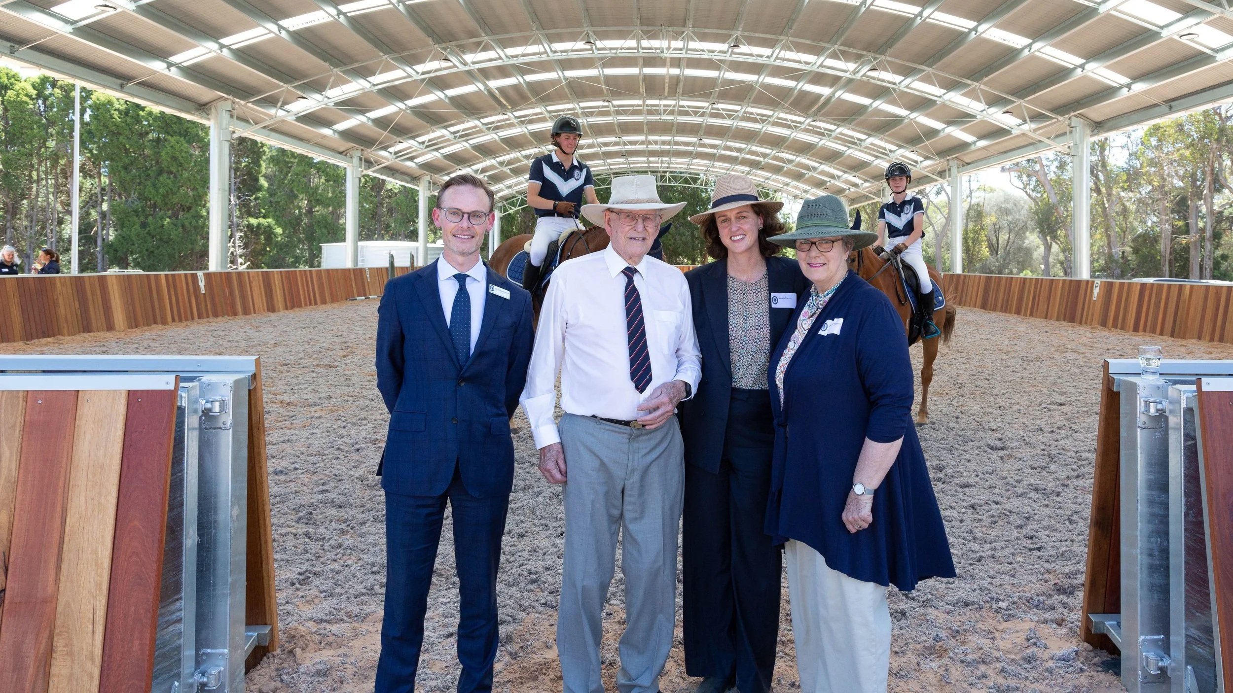 Group of four adults standing in front of two children riding horses in an outdoor riding arena with a covered roof and green trees in the background.