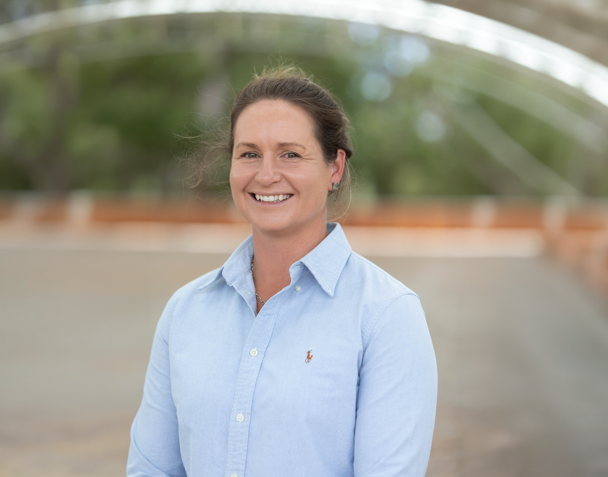 A woman with brown hair tied back, wearing a light blue button-up shirt with a small logo on the chest, smiling outdoors with blurred green trees in the background.