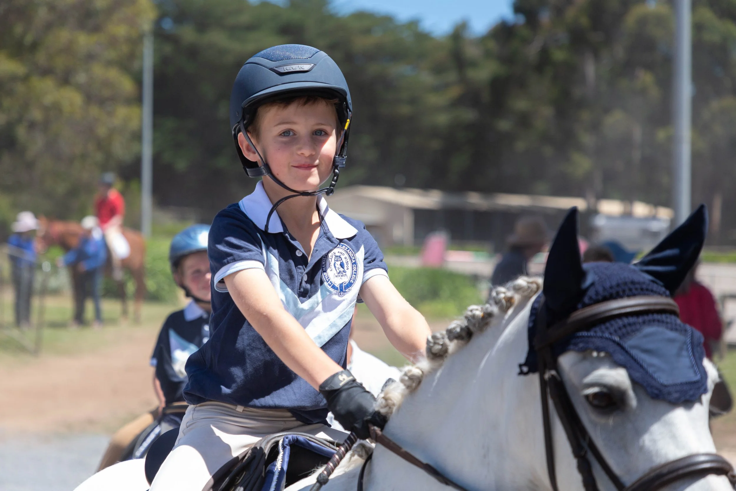 Young boy wearing a helmet and riding a horse during a daytime outdoor horse riding event with other riders and horses visible in the background.