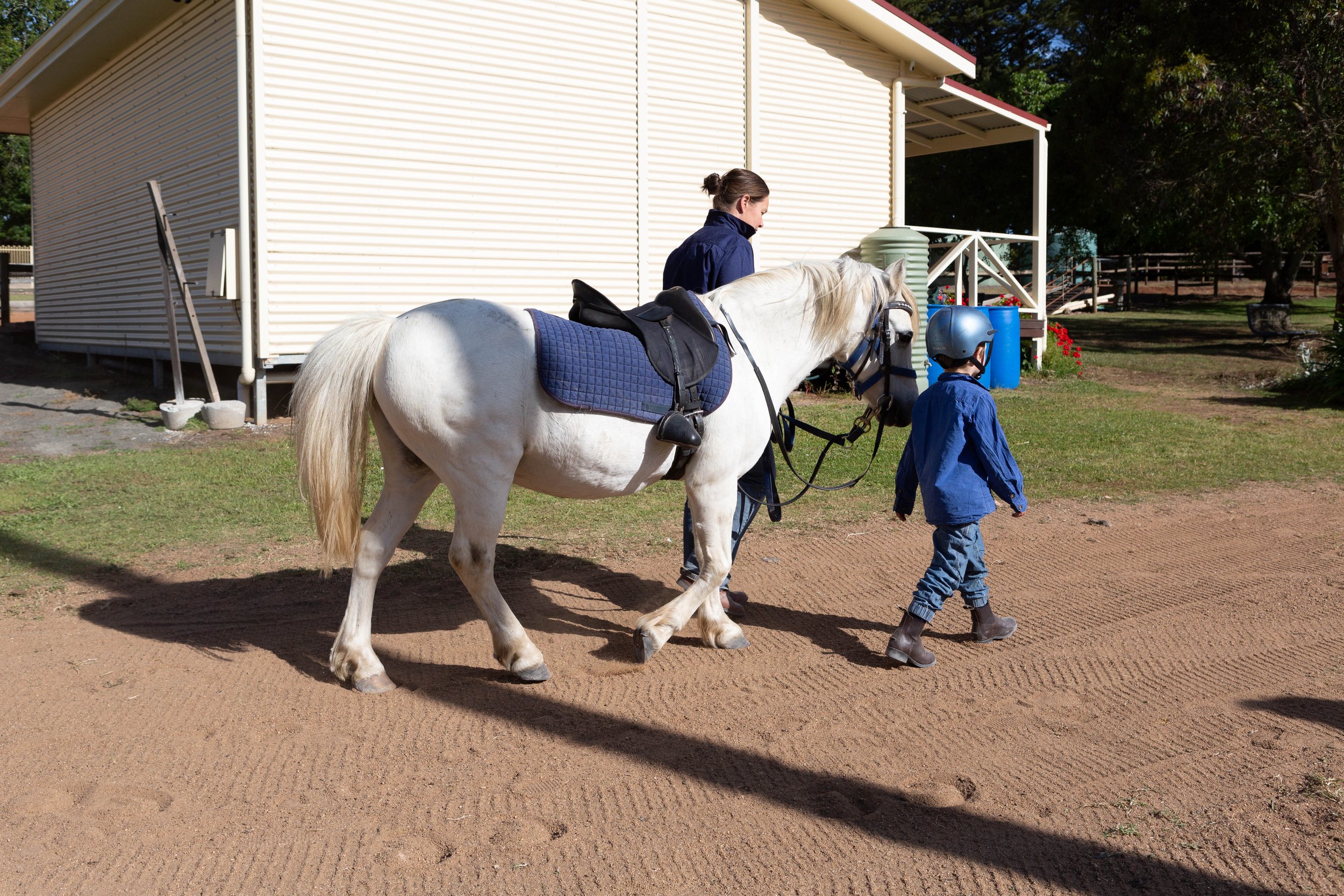 A woman and a young boy walk a white horse outside a building with tan siding and a porch. The woman is holding the horse's reins, and the boy is walking beside them. The horse is saddled and bridled.