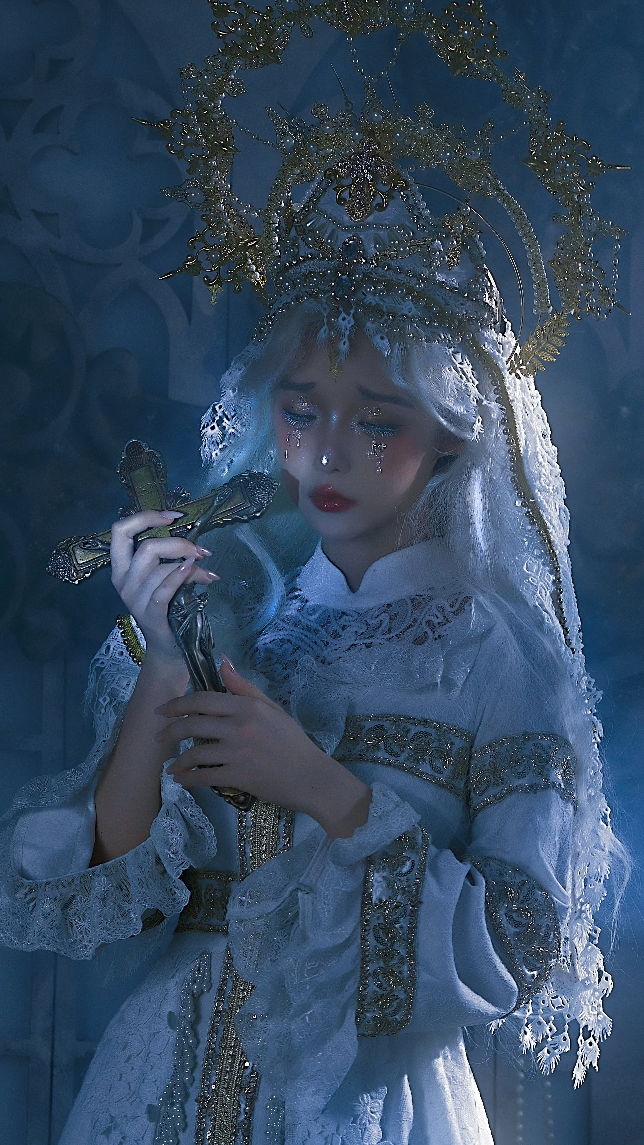 A woman in elaborate white and gold lace dress holding a crucifix, with a lace veil and ornate gold headpiece, tears on her face, in a dim, mystical setting.