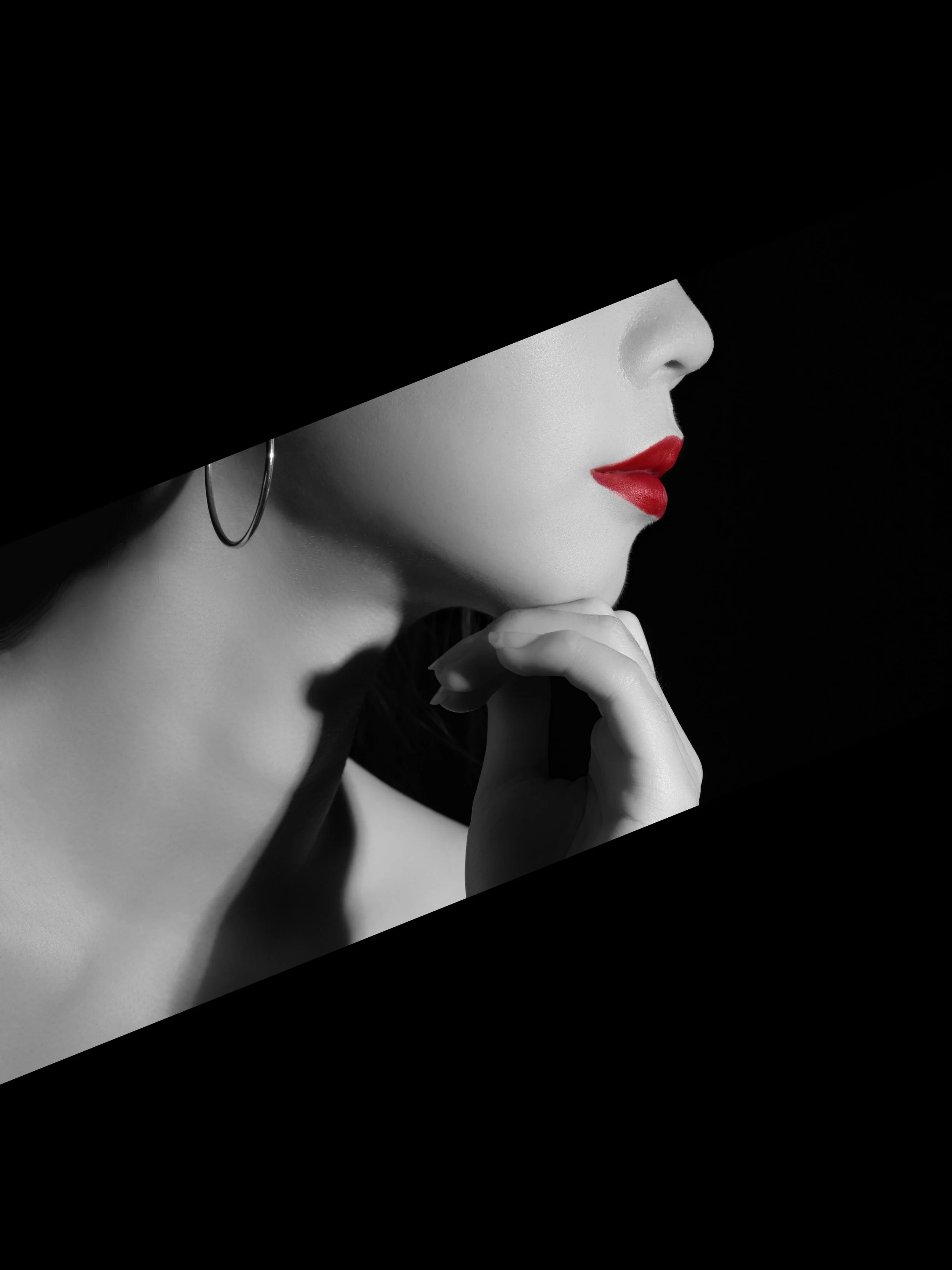 Close-up of a woman’s face, showing red lipstick, wearing a hat covering her eyes, with her hand near her chin, in black and white with the women's lips being the only color.