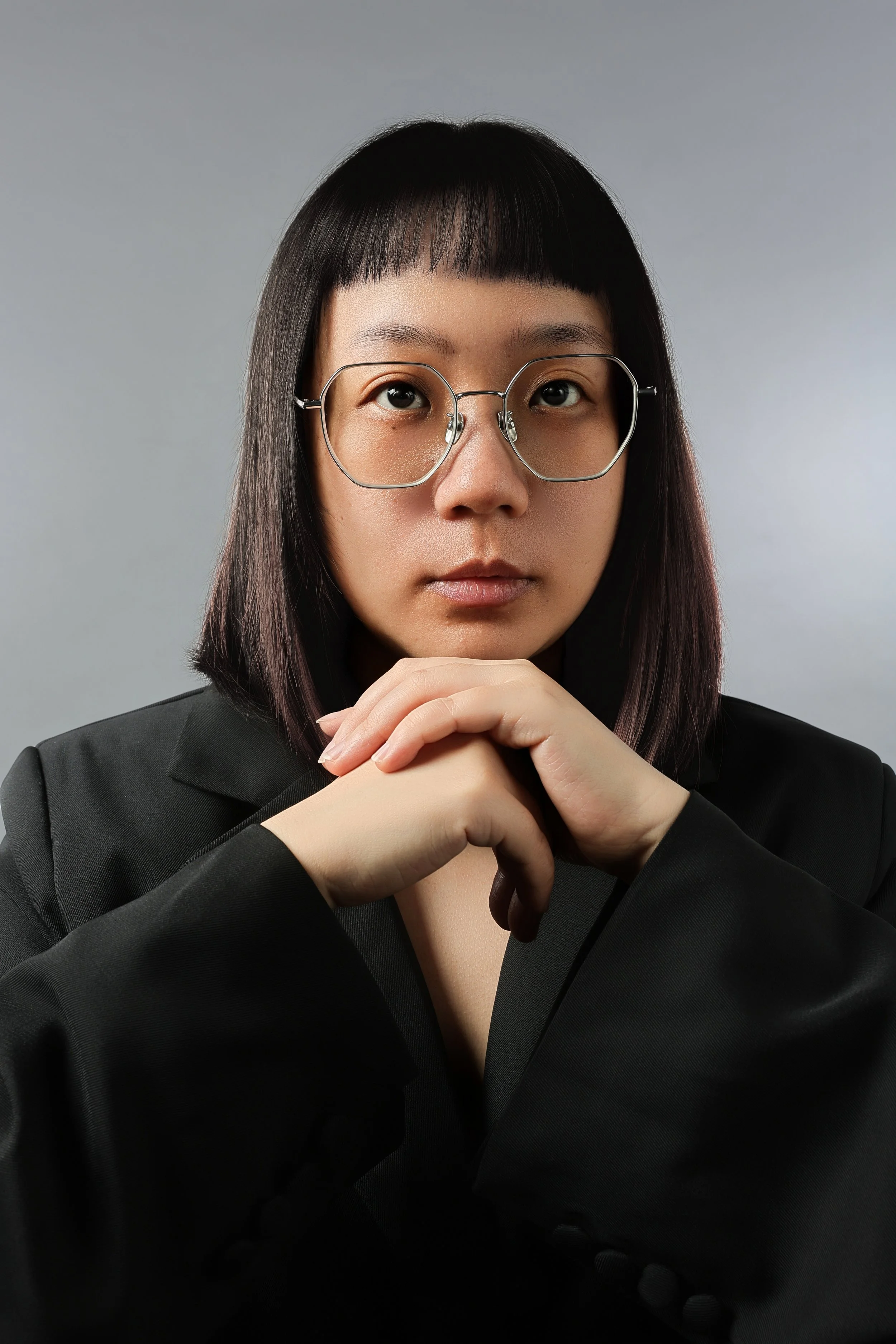 Portrait of a woman with glasses, wearing a black blazer, with her hands clasped in front of her face, against a plain background.
