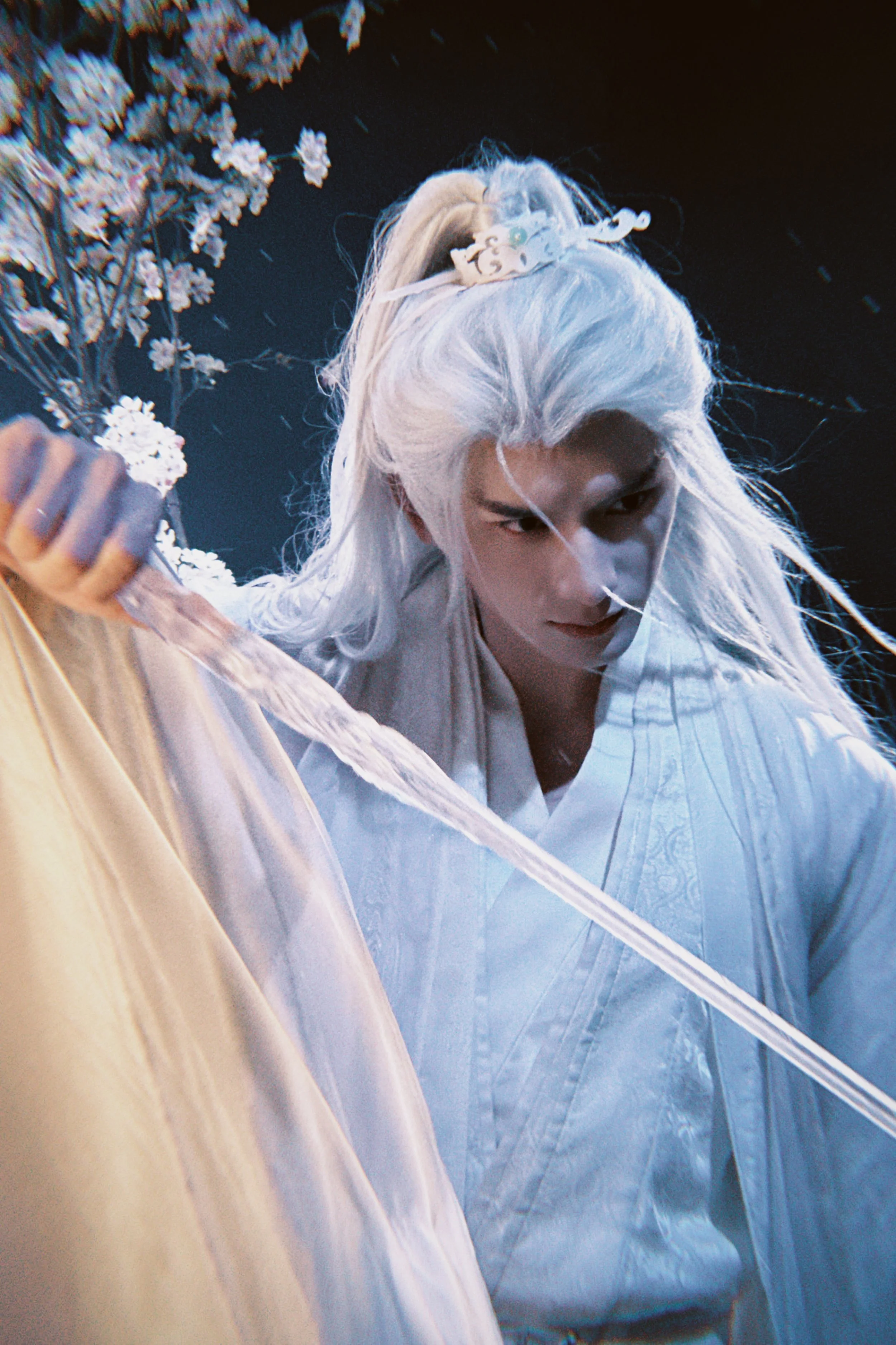 A person with long white hair dressed in traditional white clothing, holding a sword, with flowers and falling petals in background.