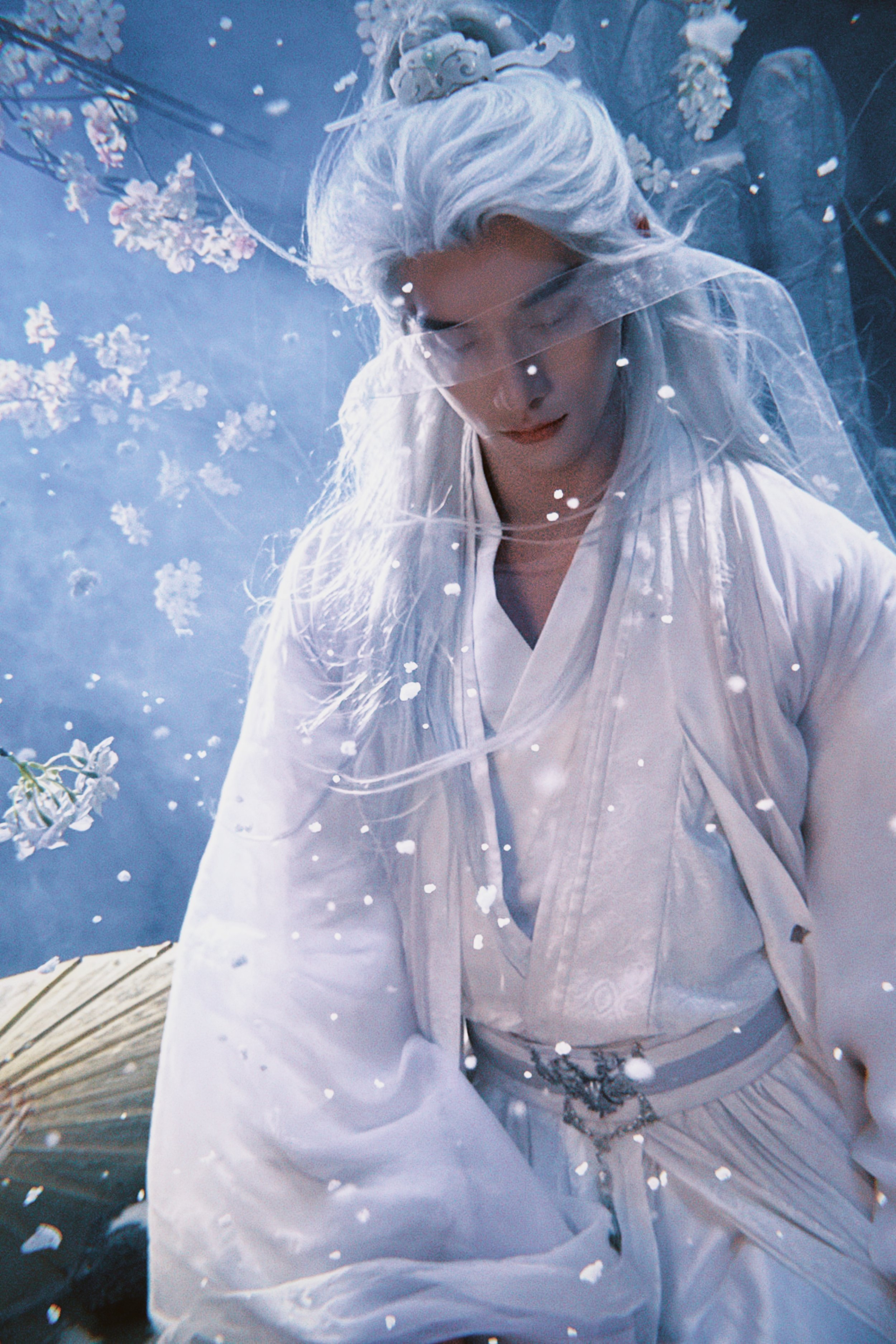 A person dressed as a fantasy character with long, flowing white hair, wearing white robes, standing behind a glass surface with snowflake and flower patterns, creating a mystical scene.