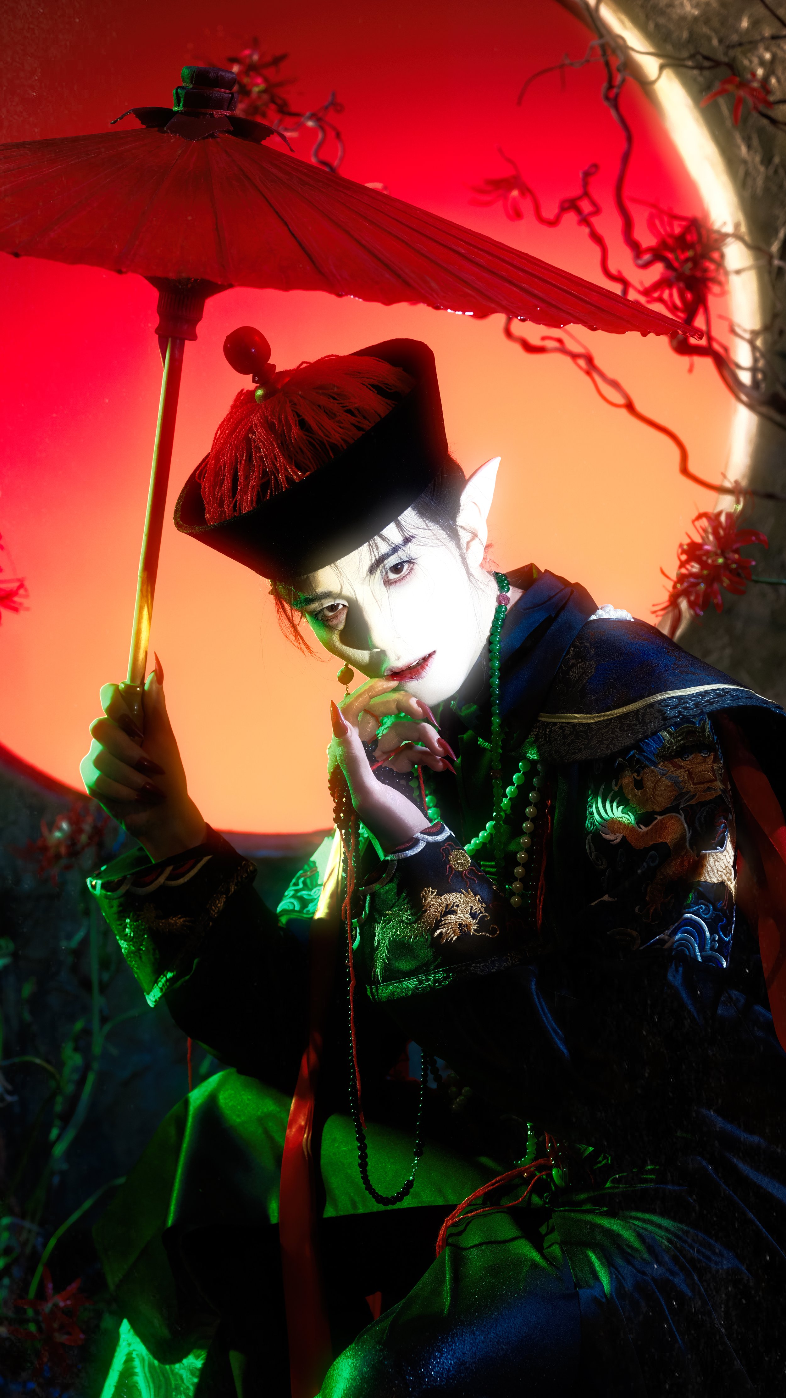 A person dressed as a fantasy elf or vampire with pointed ears, wearing a traditional Chinese embroidered robe, and accessories, holding a red parasol under colorful lighting, with a vivid red and green background.
