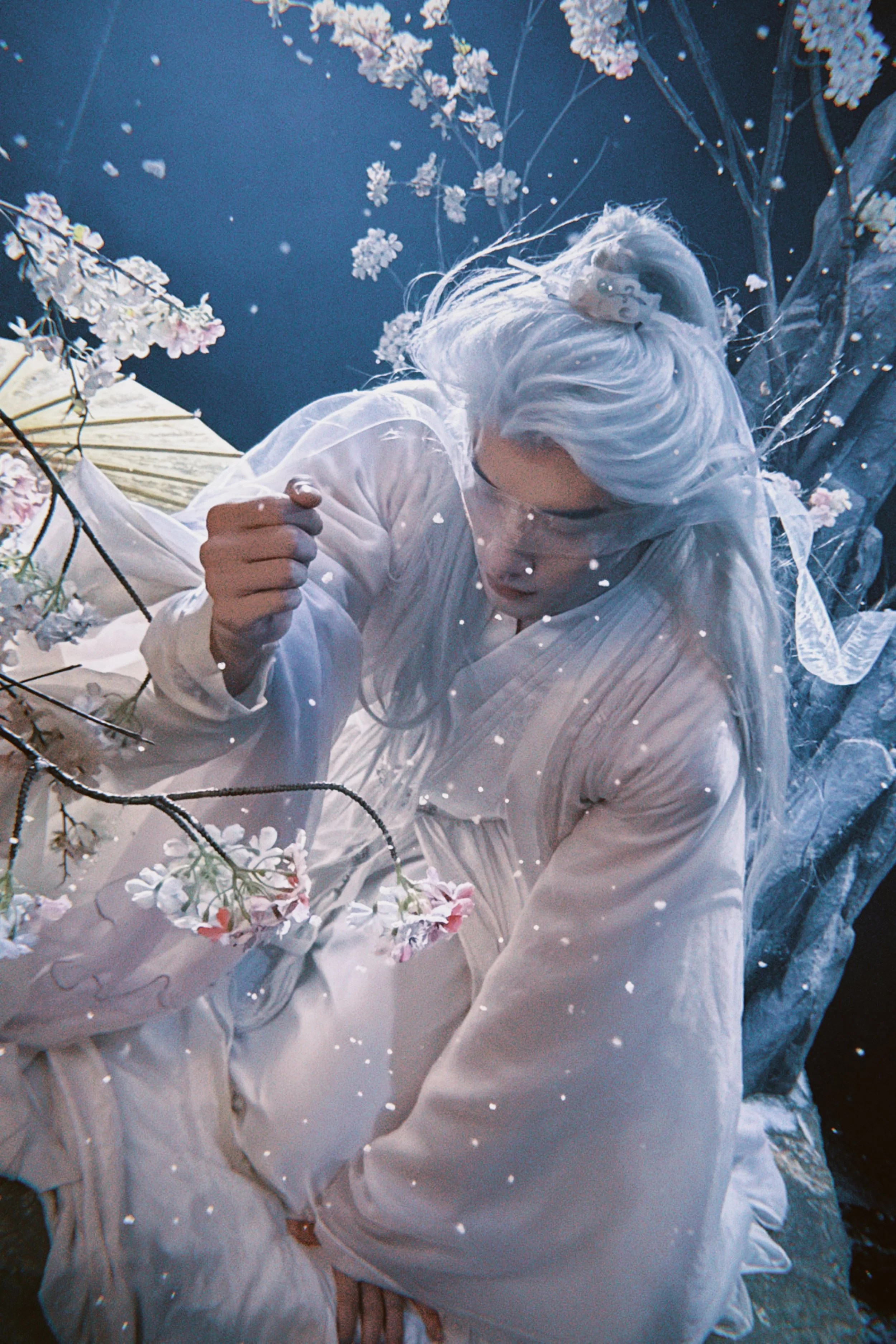 A person with long white hair dressed in white, surrounded by pink cherry blossoms, in a dreamy, soft-lit scene.