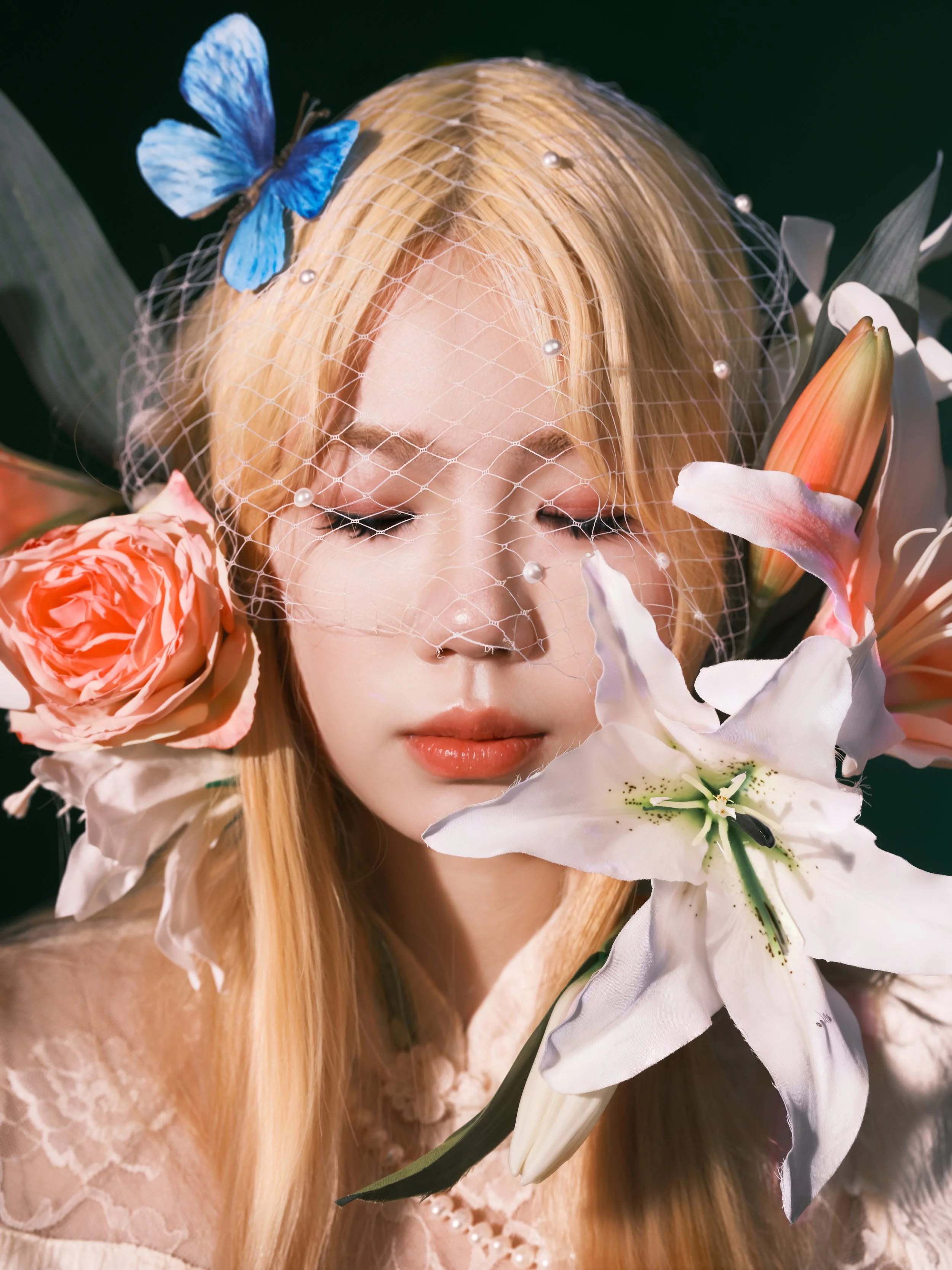 A woman with long blonde hair and eye makeup features holds a large white lily flower near her face, surrounded by pink roses, a peach lily, a blue butterfly, and a pearl-embellished white net veil.