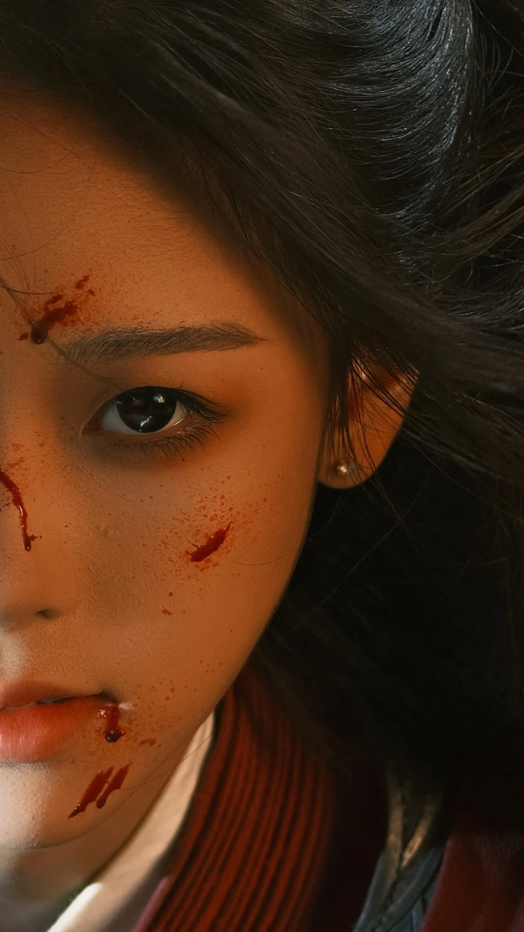 A close-up of a young woman's face with fake blood and wounds, showing her eye, eyebrow, part of her nose, and lips, with dark hair and a small earring.