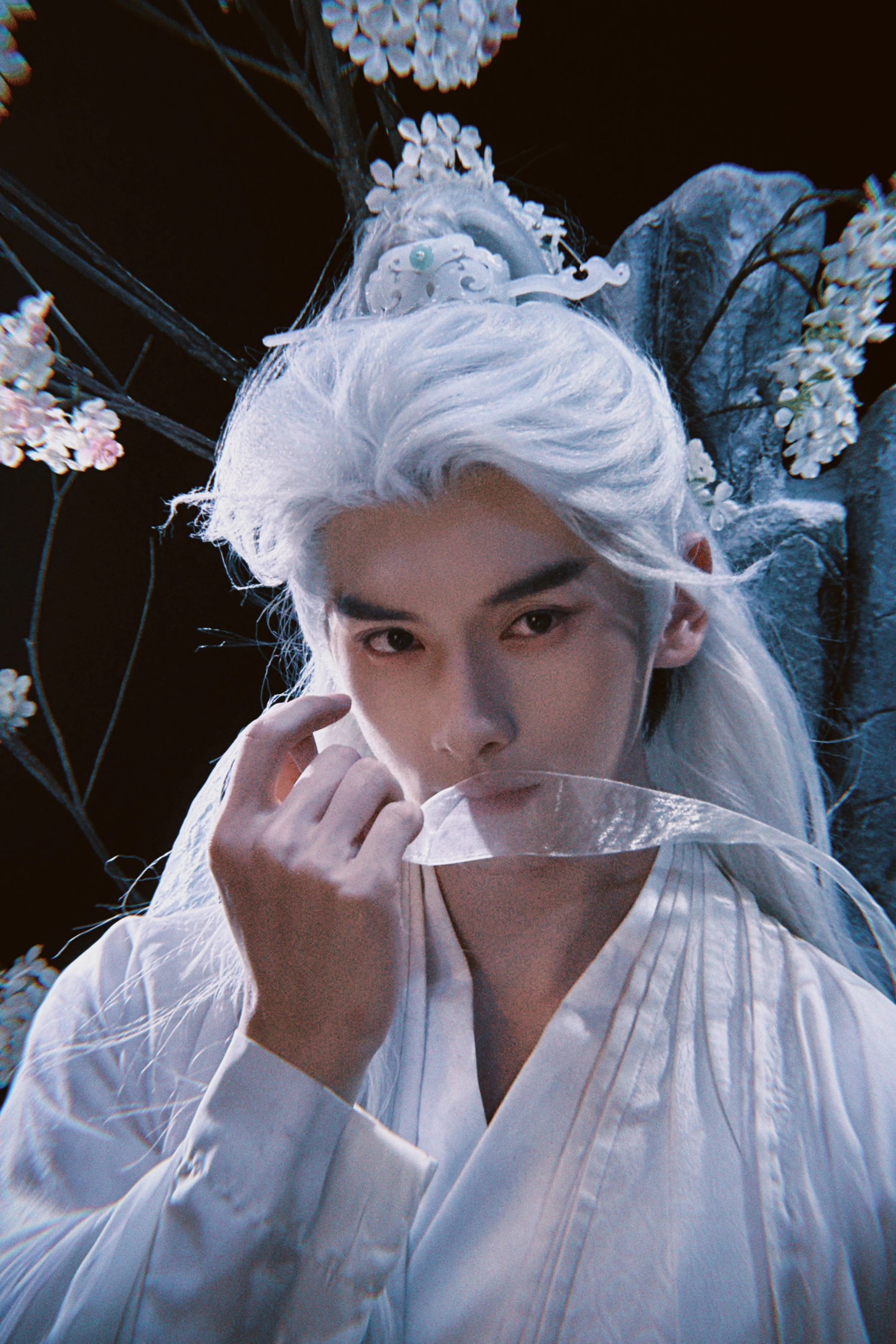 A person with long white hair holding a transparent leaf mask near their lips, dressed in white, with a dark background and white flowers surrounding.