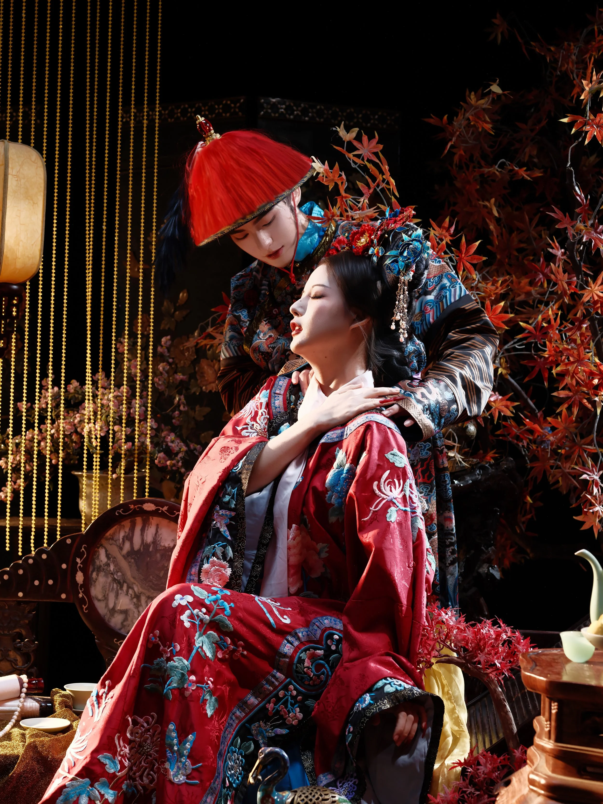 Two women in traditional Asian attire, with one woman seated and the other leaning over her, in a decorated setting with red leaves and hanging gold ornaments.