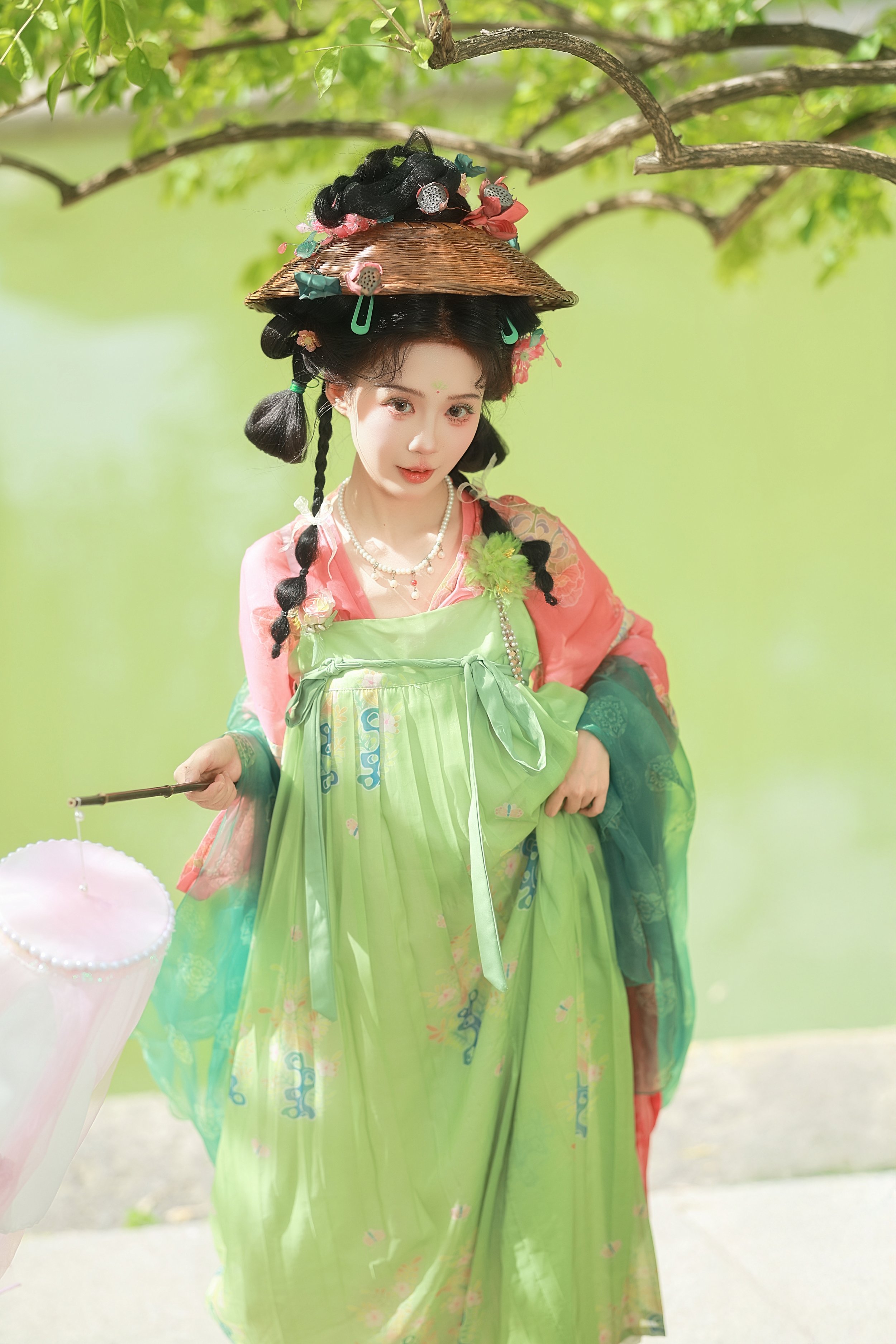 A woman dressed in traditional Asian attire with a green and pink kimono, holding a pink lantern, standing outdoors under a tree with green leaves.