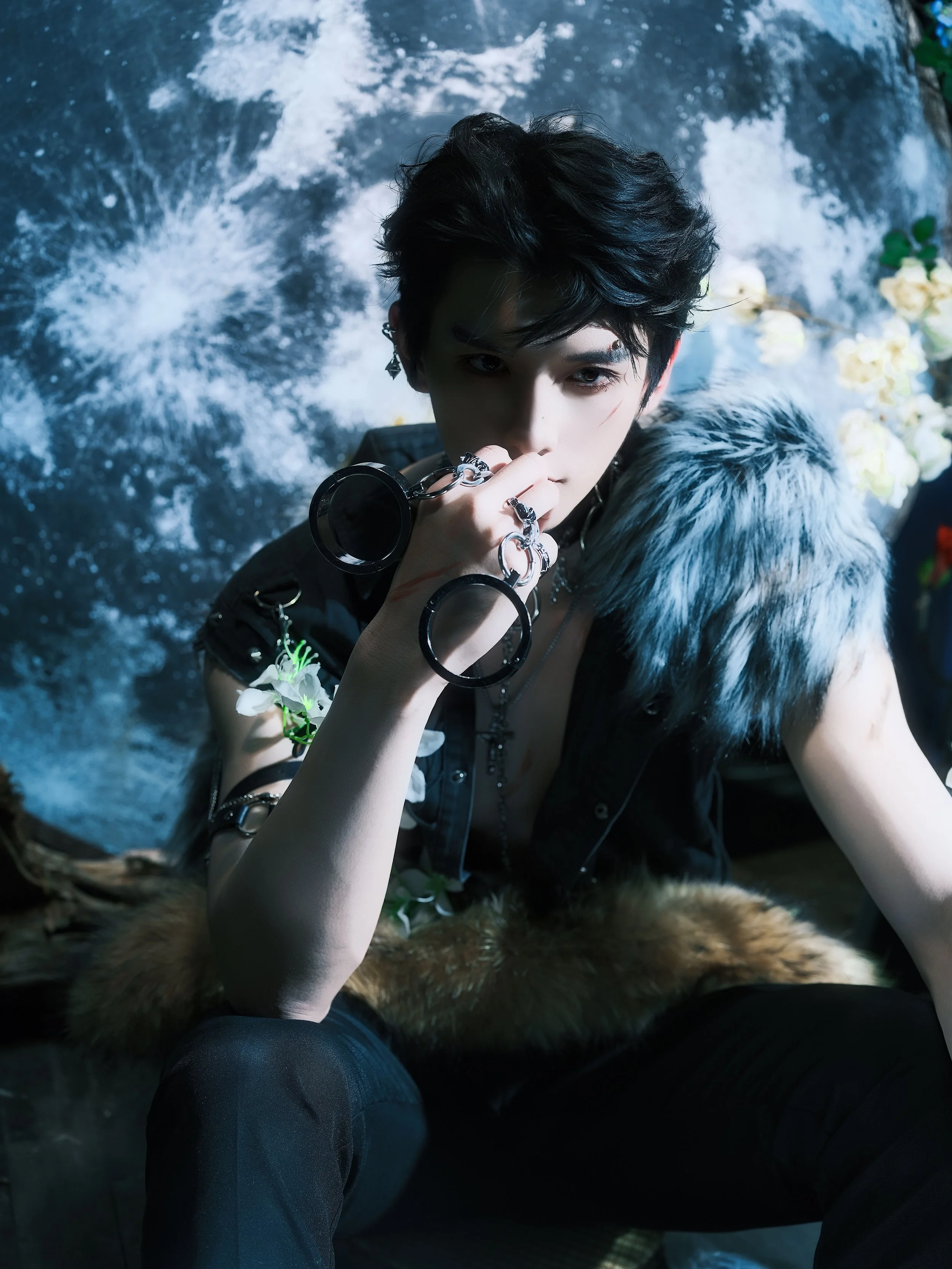 A young man with dark, tousled hair and multiple rings on his fingers, wearing a black sleeveless outfit with a large gray fur shoulder piece, sitting against a backdrop of a blue water scene with white foam and some flowers in the background.
