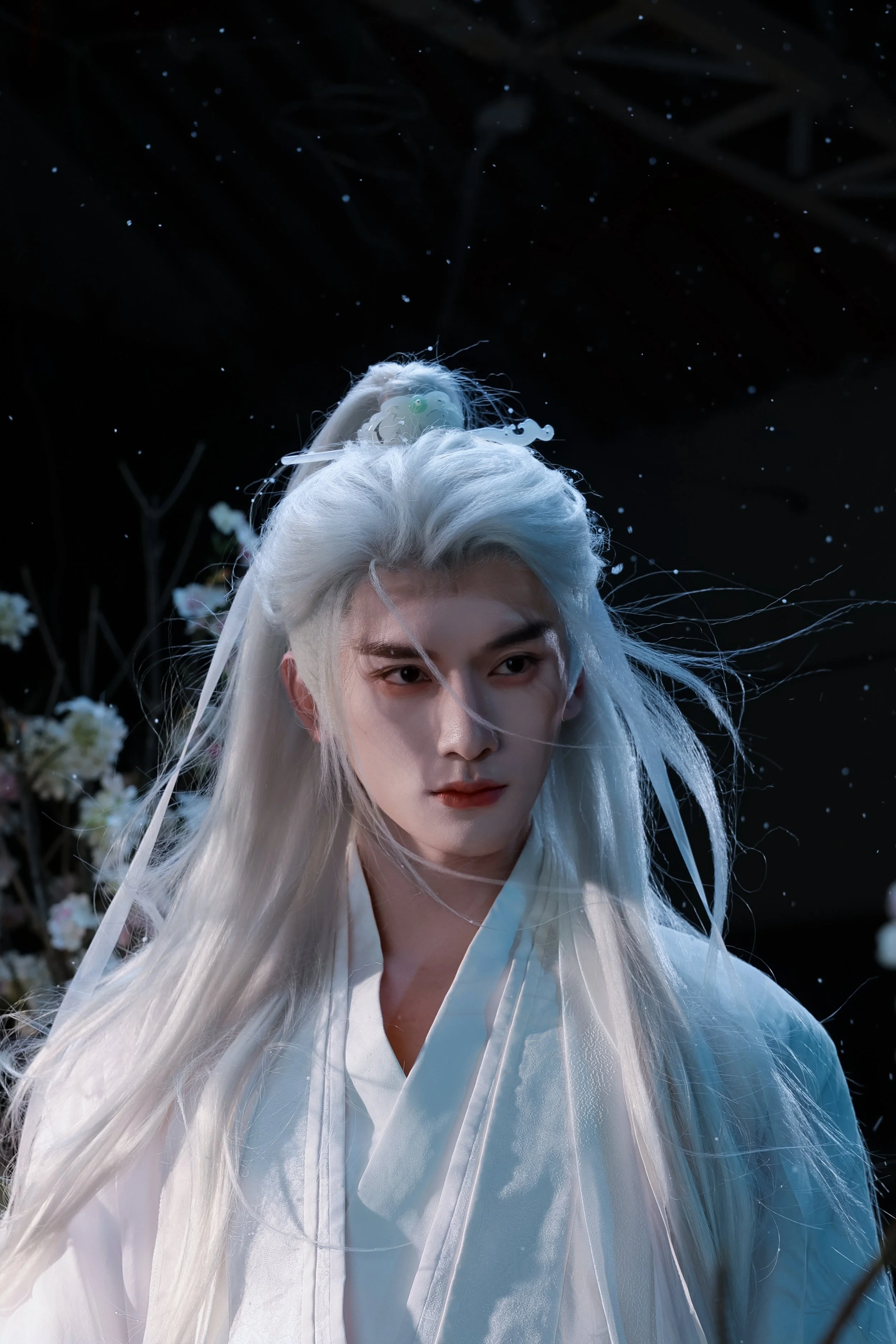 Person with long white hair wearing traditional white clothing, with wind blowing strands of hair, standing amid white flowers with a dark background and falling snow or petals.