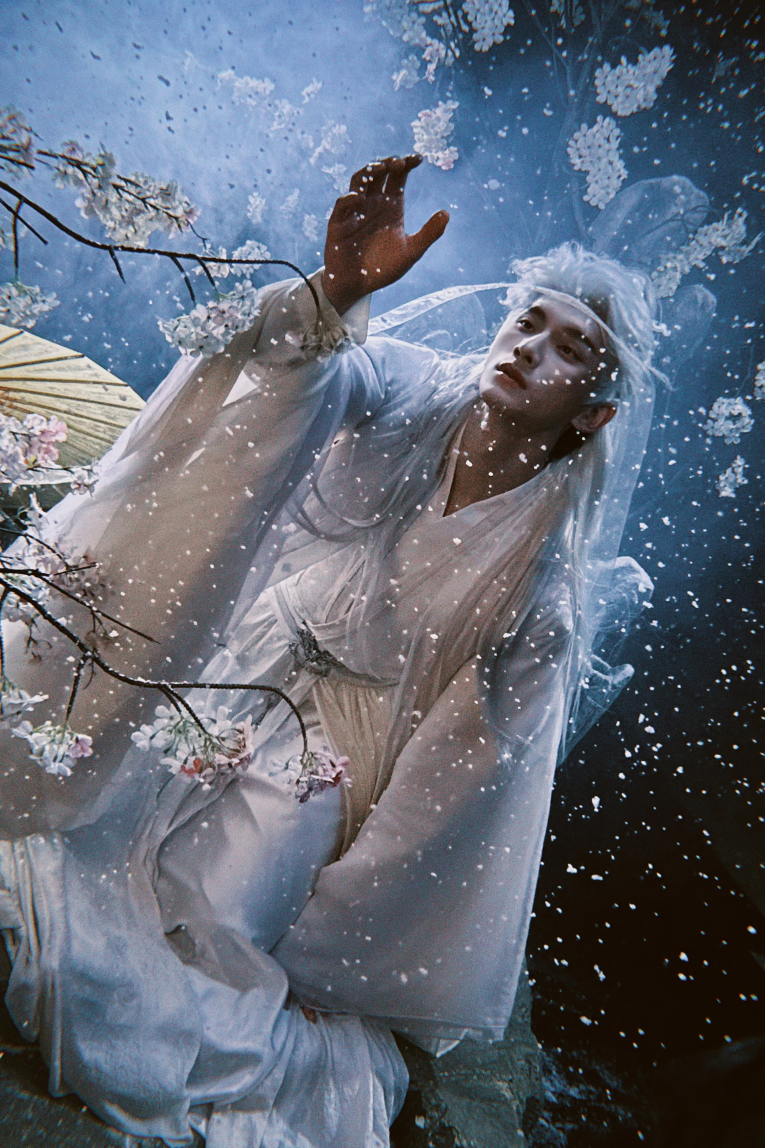 A person with white hair and makeup dressed in white, reaching out underwater amidst floating flowers and branches.