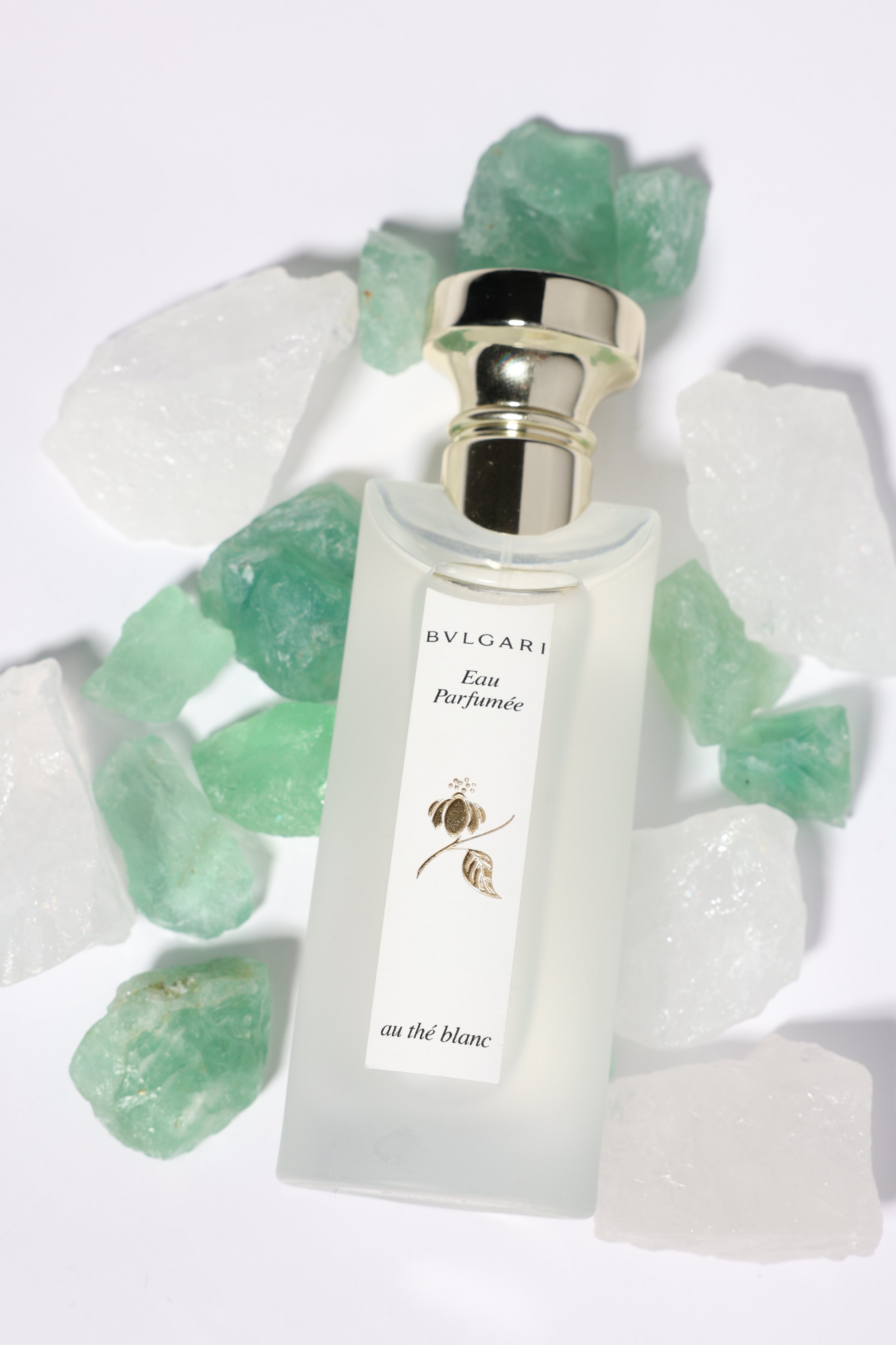 A frosted perfume bottle labeled BVLGARI Eau Parfumée au thé blanc surrounded by green and white crystal stones on a white background.
