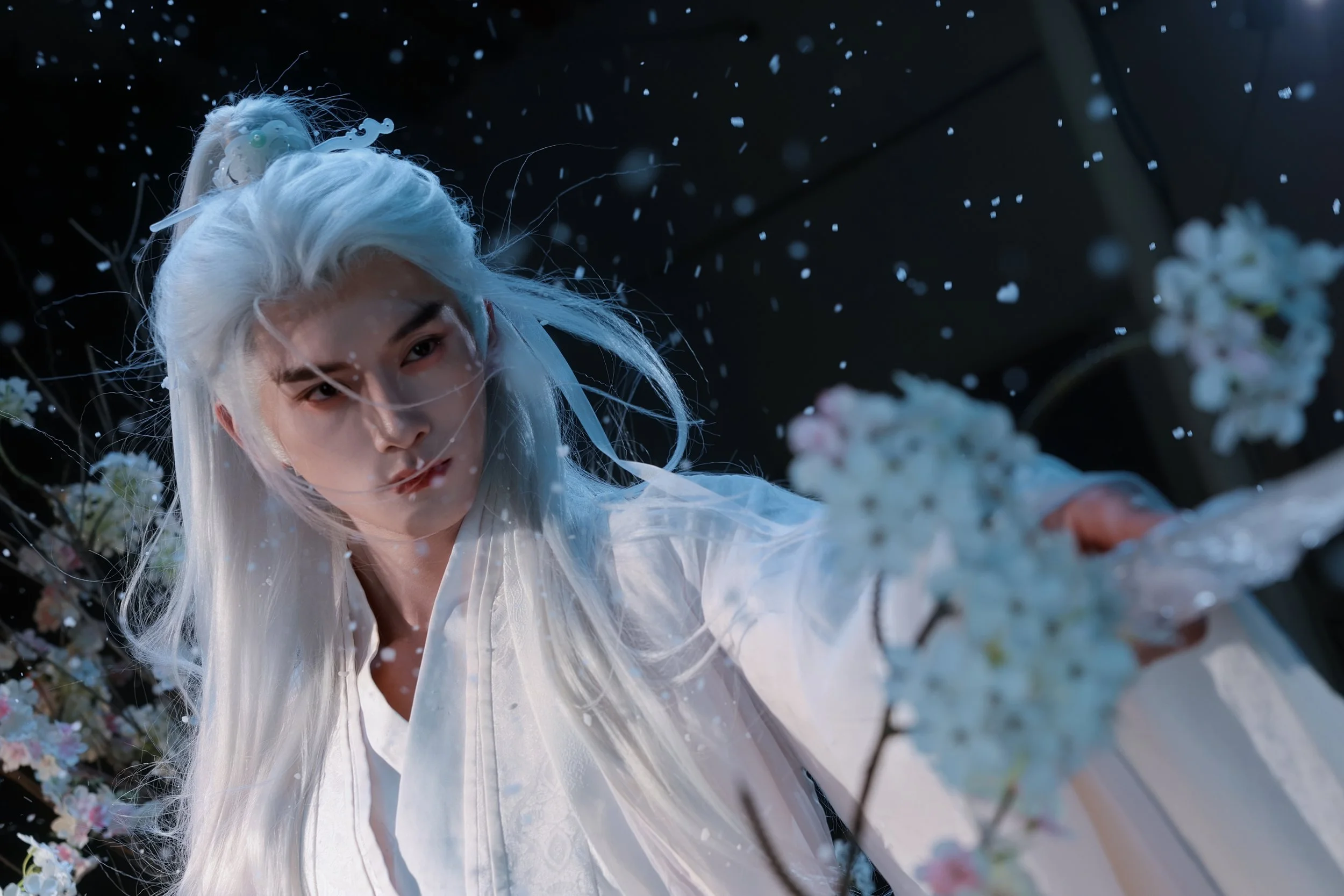 A person dressed in white with long white hair, surrounded by falling petals and flowers.