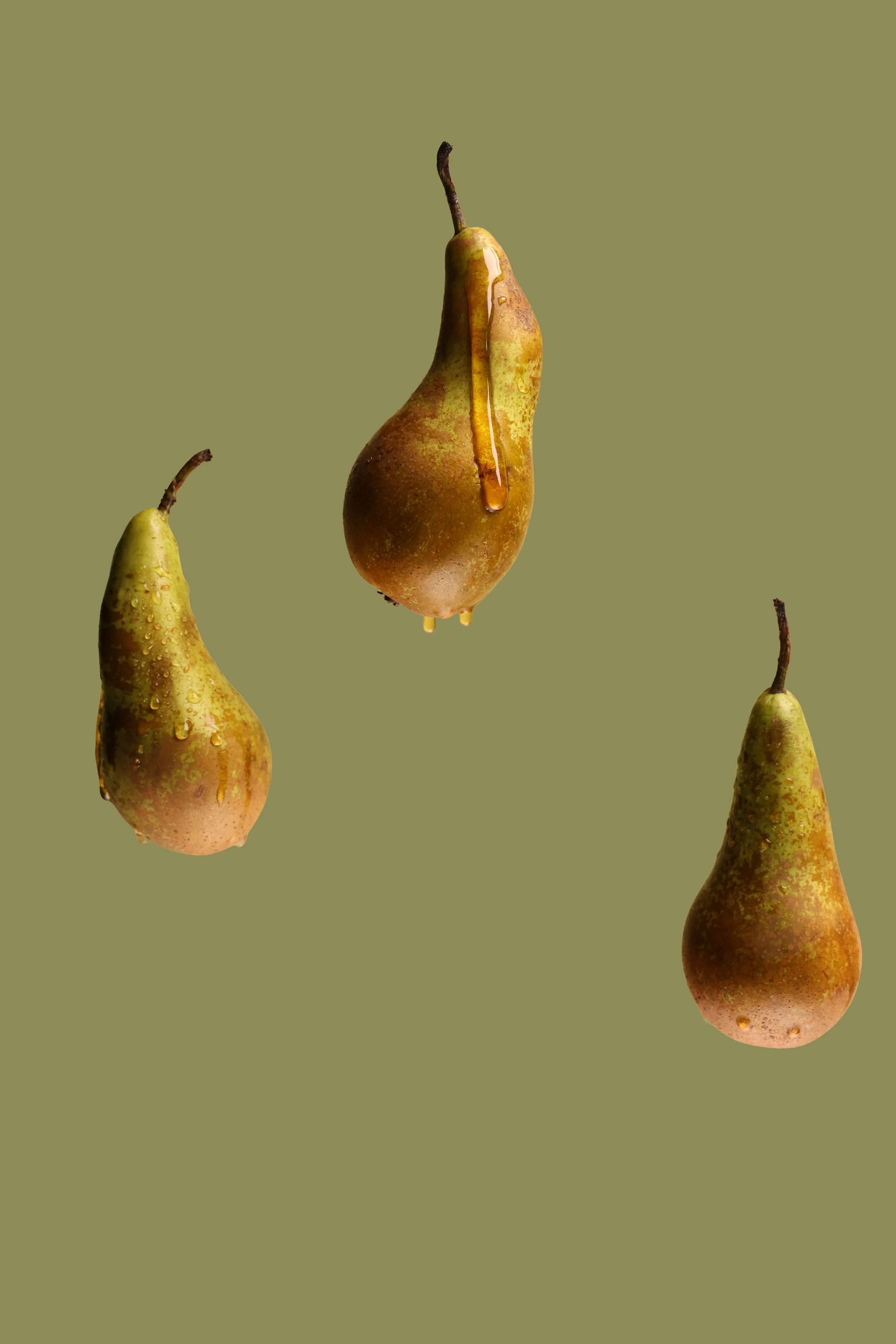 Three pears against a plain green background, one with honey dripping down and droplets of water on its surface.