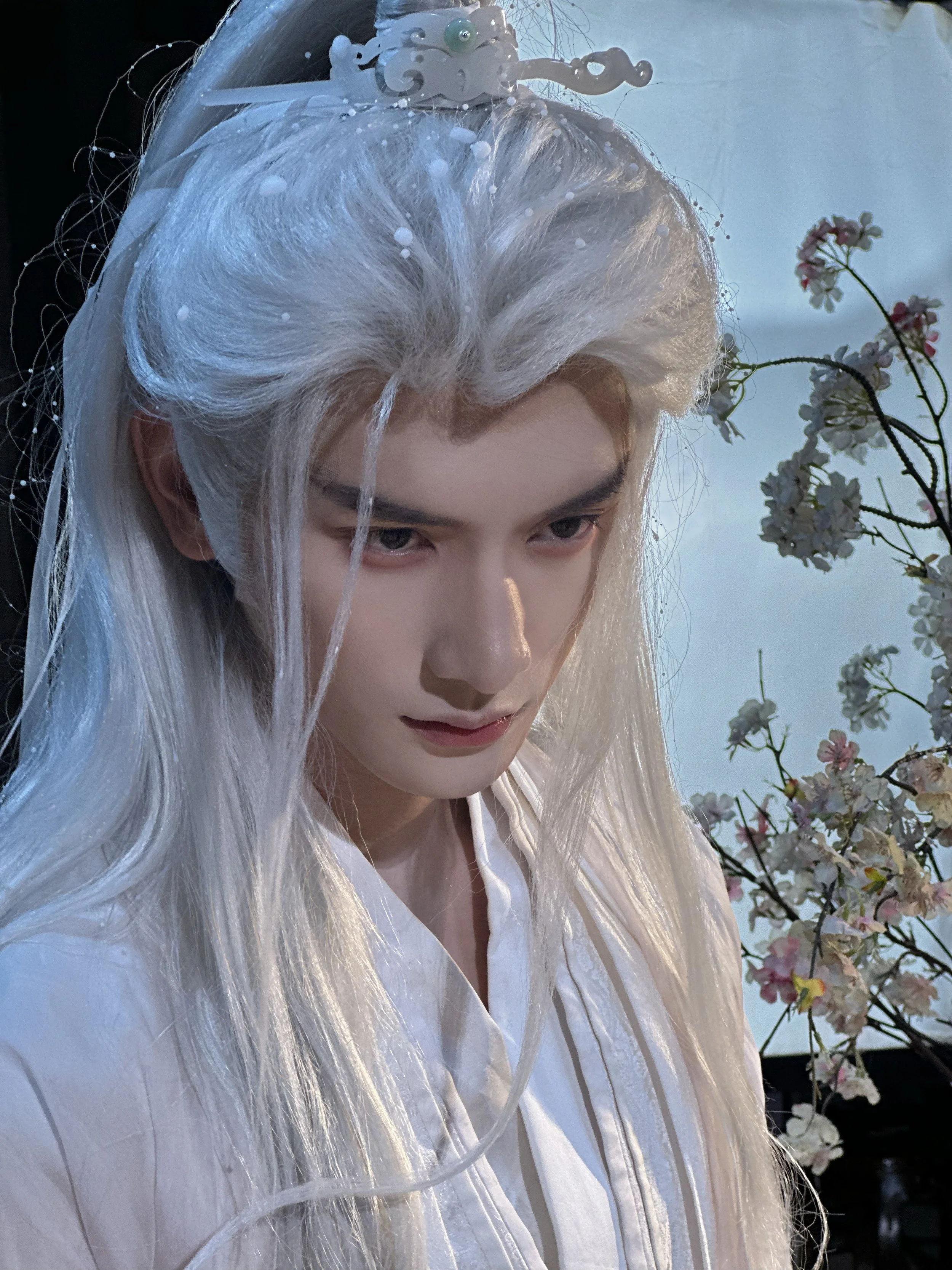 A person with long, white hair styled with a pearl and floral hair accessory, wearing a white top, with a backdrop of pink and white flowers.