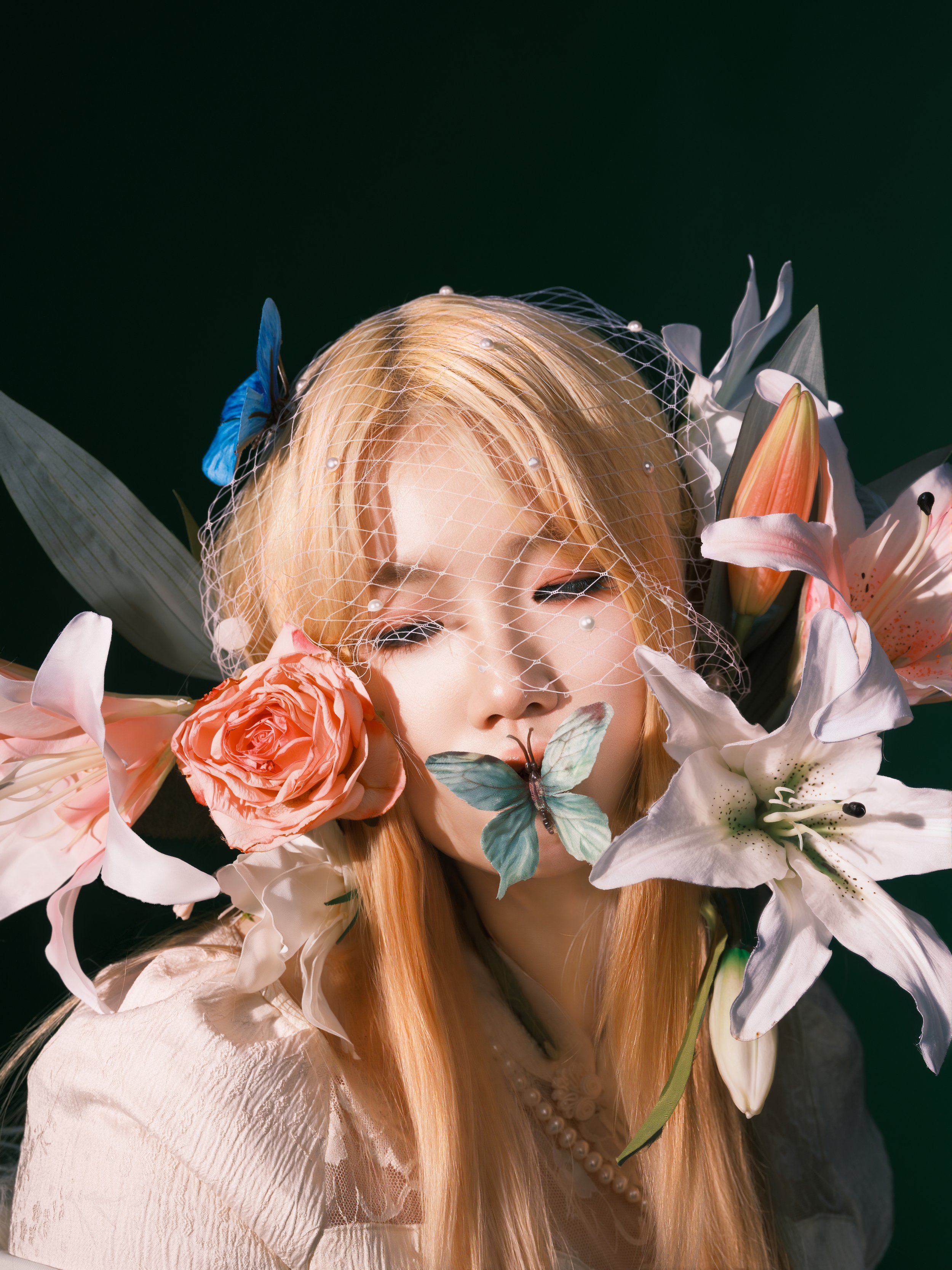 A woman with closed eyes surrounded by lilies, roses, and a butterfly on her lips, wearing a veil and pearl necklace, with a blue bow in her blonde hair.