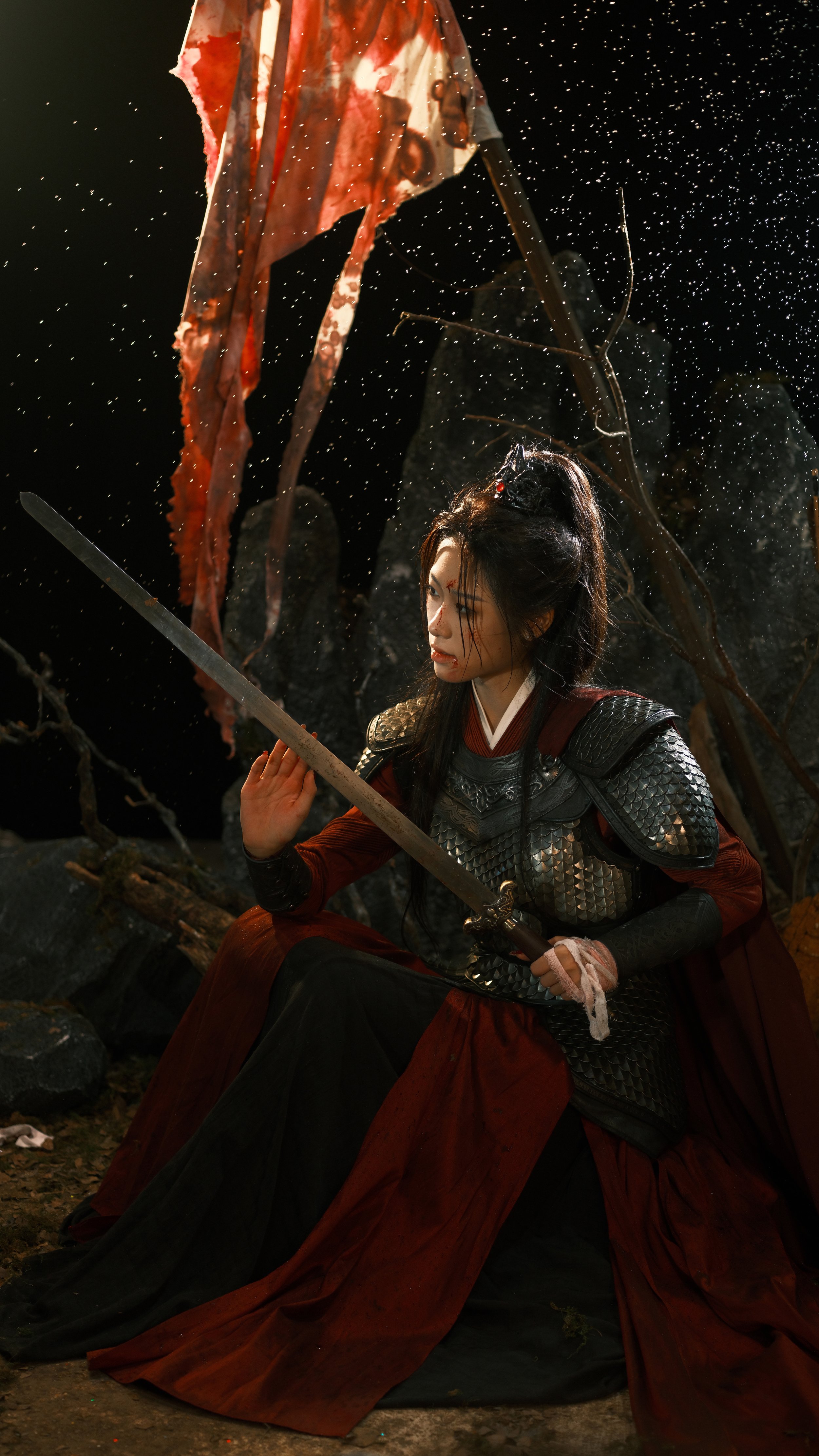 A woman dressed in medieval armor, sitting outdoors at night with a sword, surrounded by foliage and falling rain or snow, holding a sword with a serious expression.
