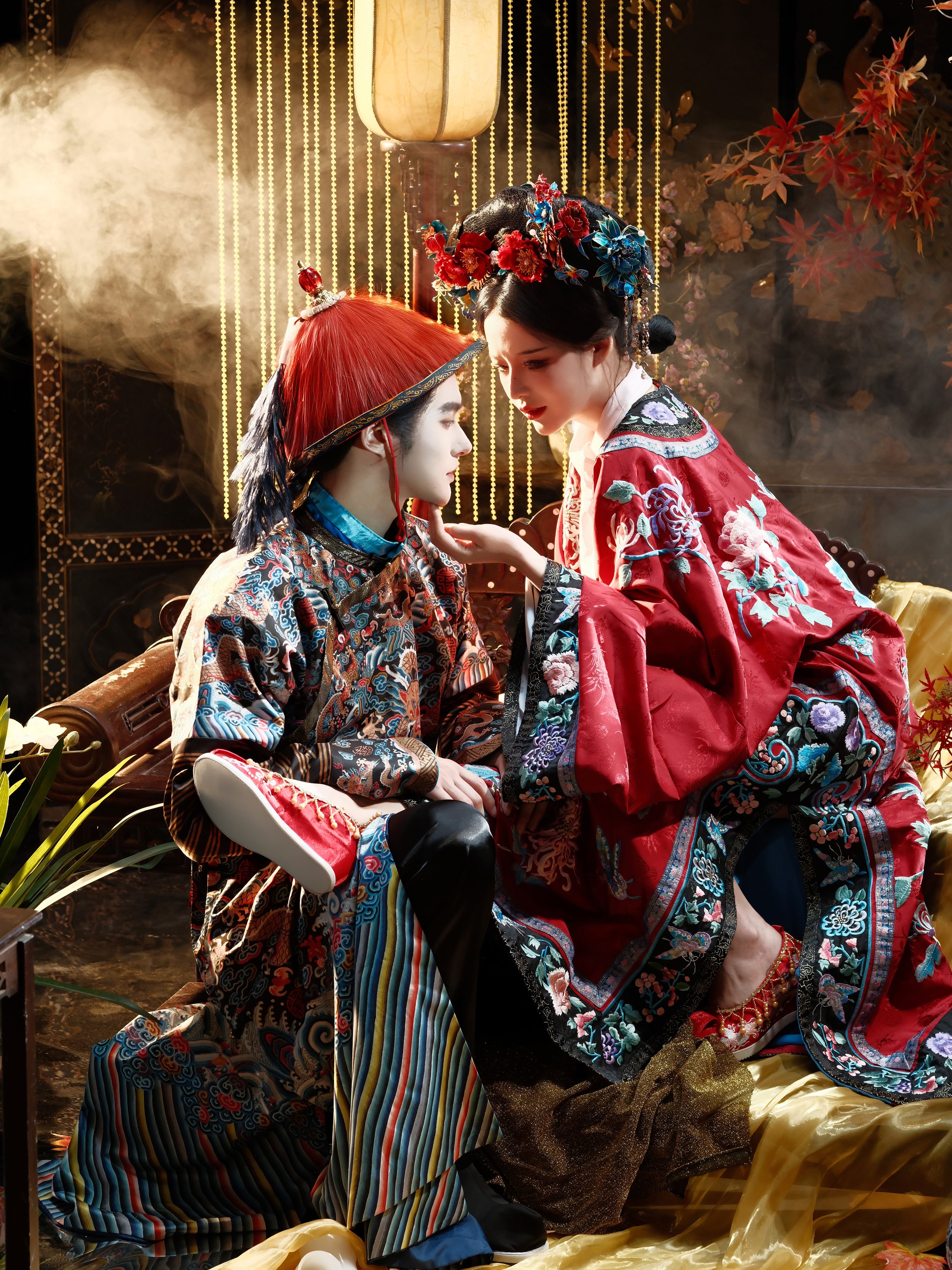 Two women in traditional Chinese attire with elaborate embroidered robes and ornate headpieces, engaged in an intimate moment indoors with warm lighting and decorative elements.