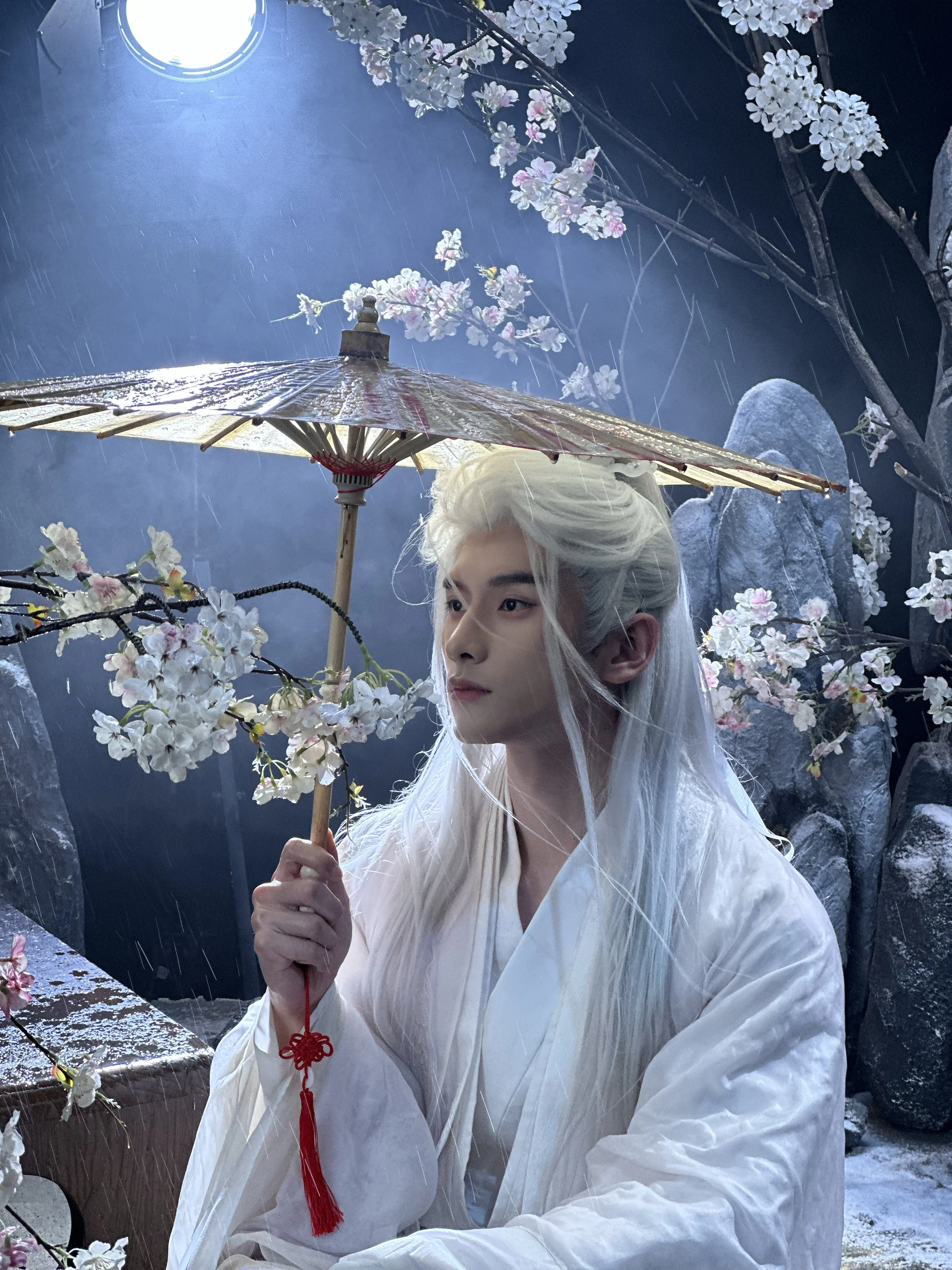 A person with long white hair dressed in traditional white clothing holding a paper umbrella decorated with cherry blossoms. The scene is set outdoors with cherry blossoms and rocks, suggesting a rainy night.