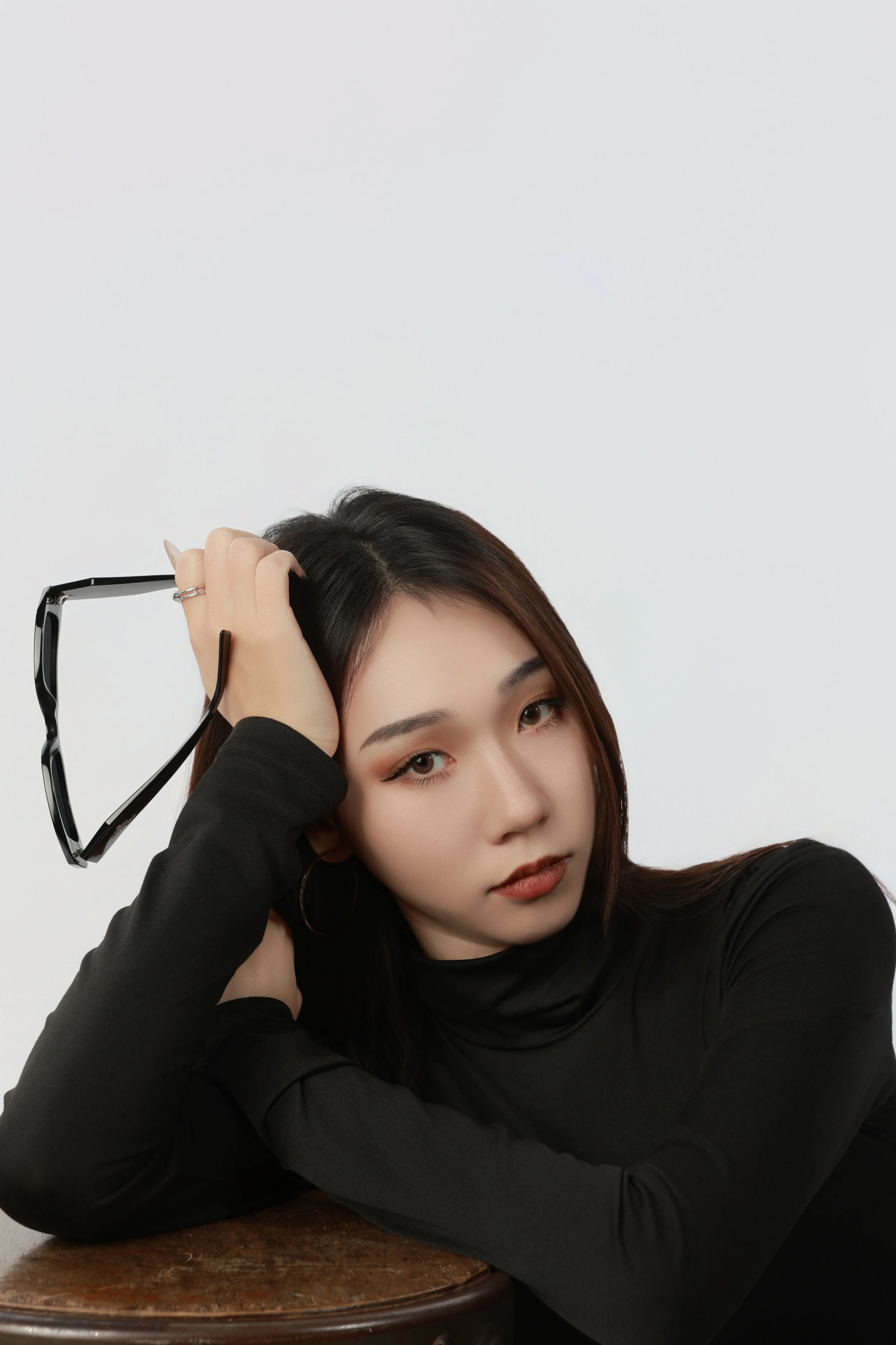 A young woman with dark hair, wearing a black turtleneck, resting her head on her hand, holding eyeglasses, sitting at a wooden table against a plain white background.