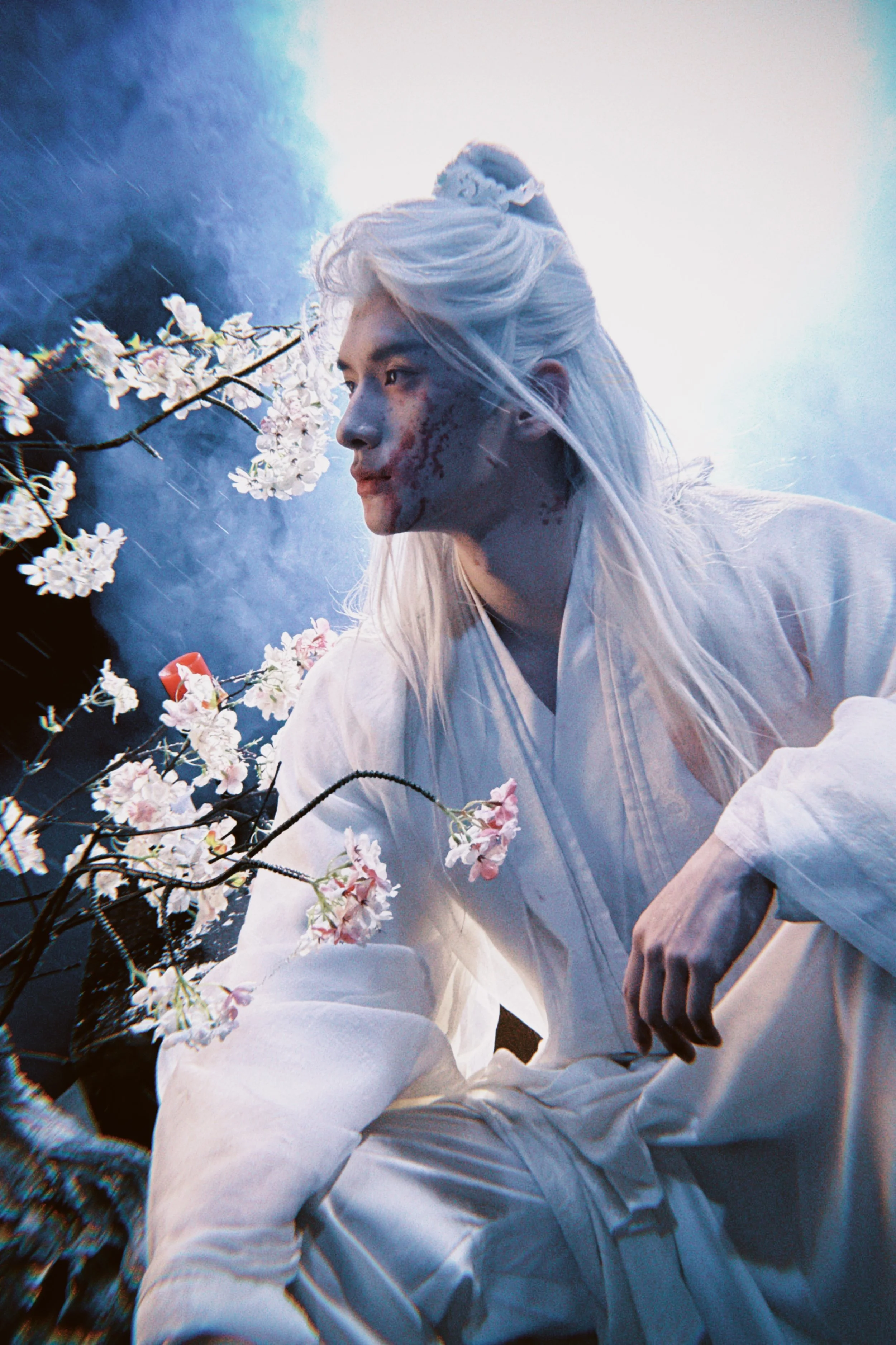 A person with long platinum blonde hair dressed in white robes, surrounded by pink cherry blossoms and vines, with a misty blue background.