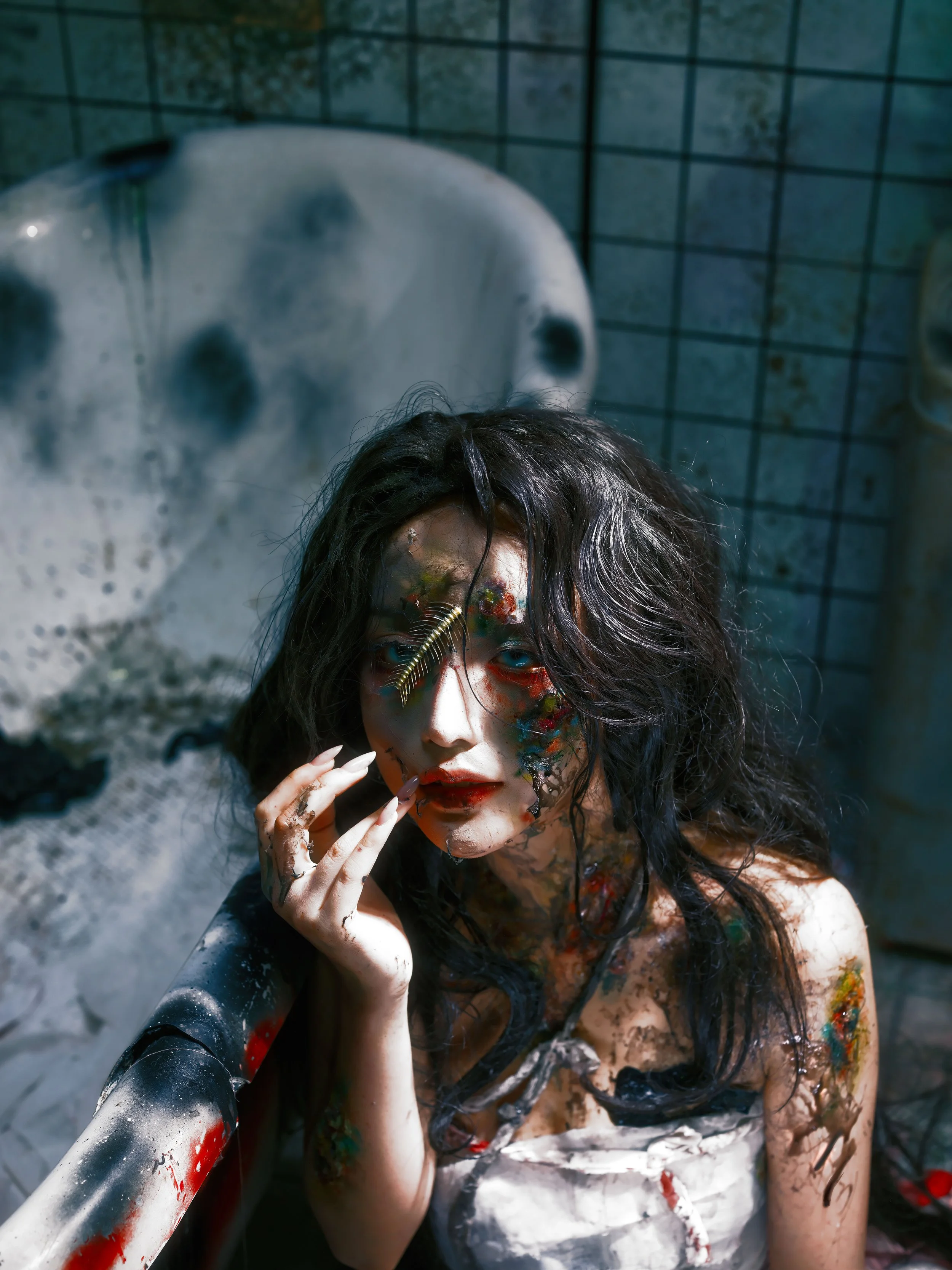 A woman with messy, dark hair and colorful, messy makeup, sitting in a dirty bathroom with graffiti on walls, surrounded by dark and eerie atmosphere.