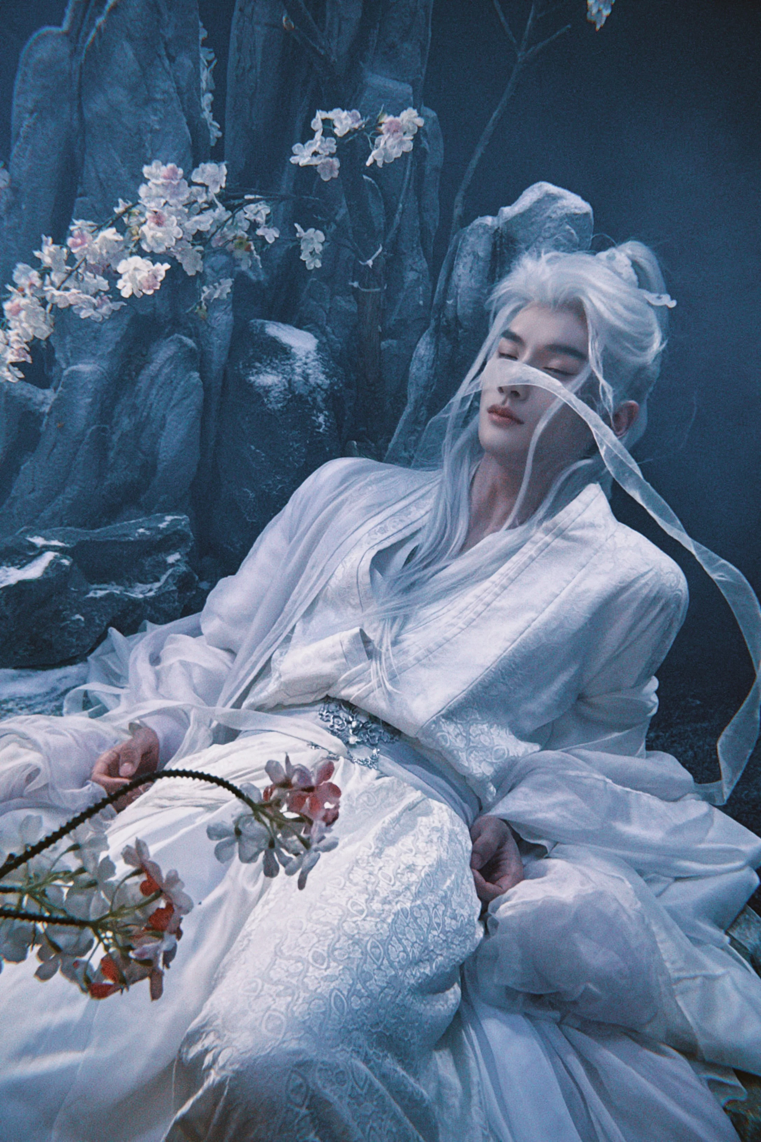 A person with long white hair, dressed in traditional white attire, is reclining against rocks with flowering branches nearby, in a stylized scene resembling a painting.