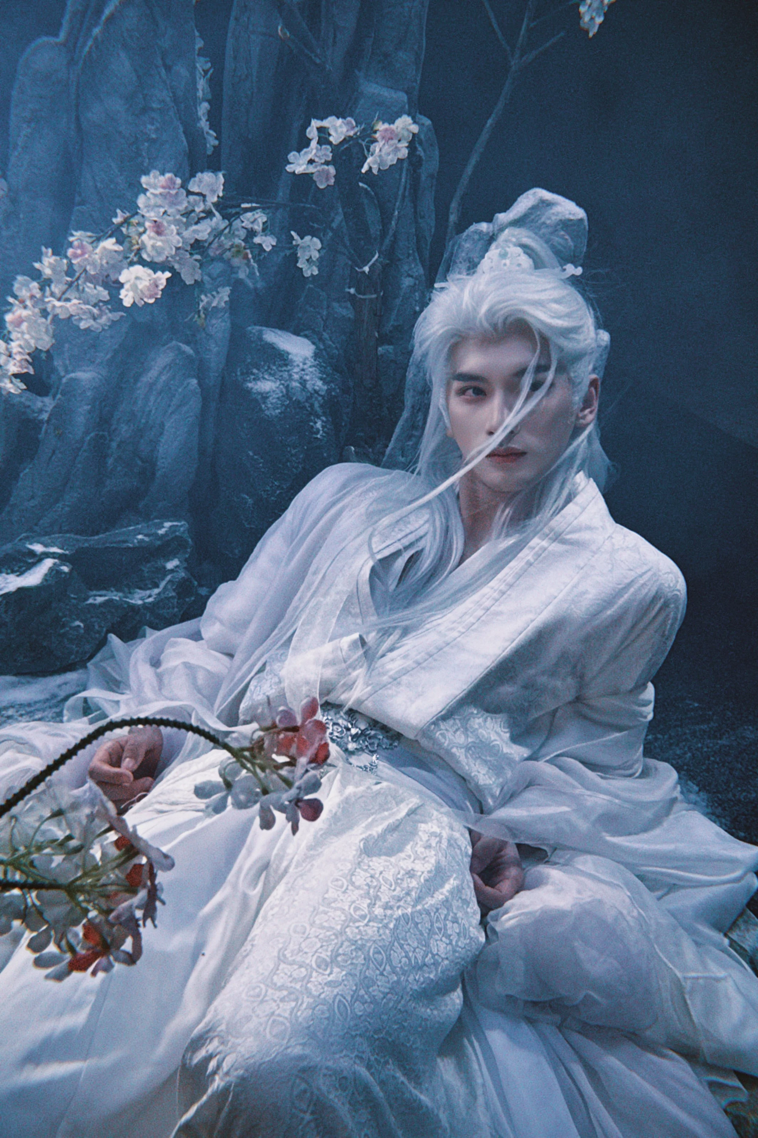 A person with long white hair in traditional white clothing, sitting against rocks with pink flowers nearby.