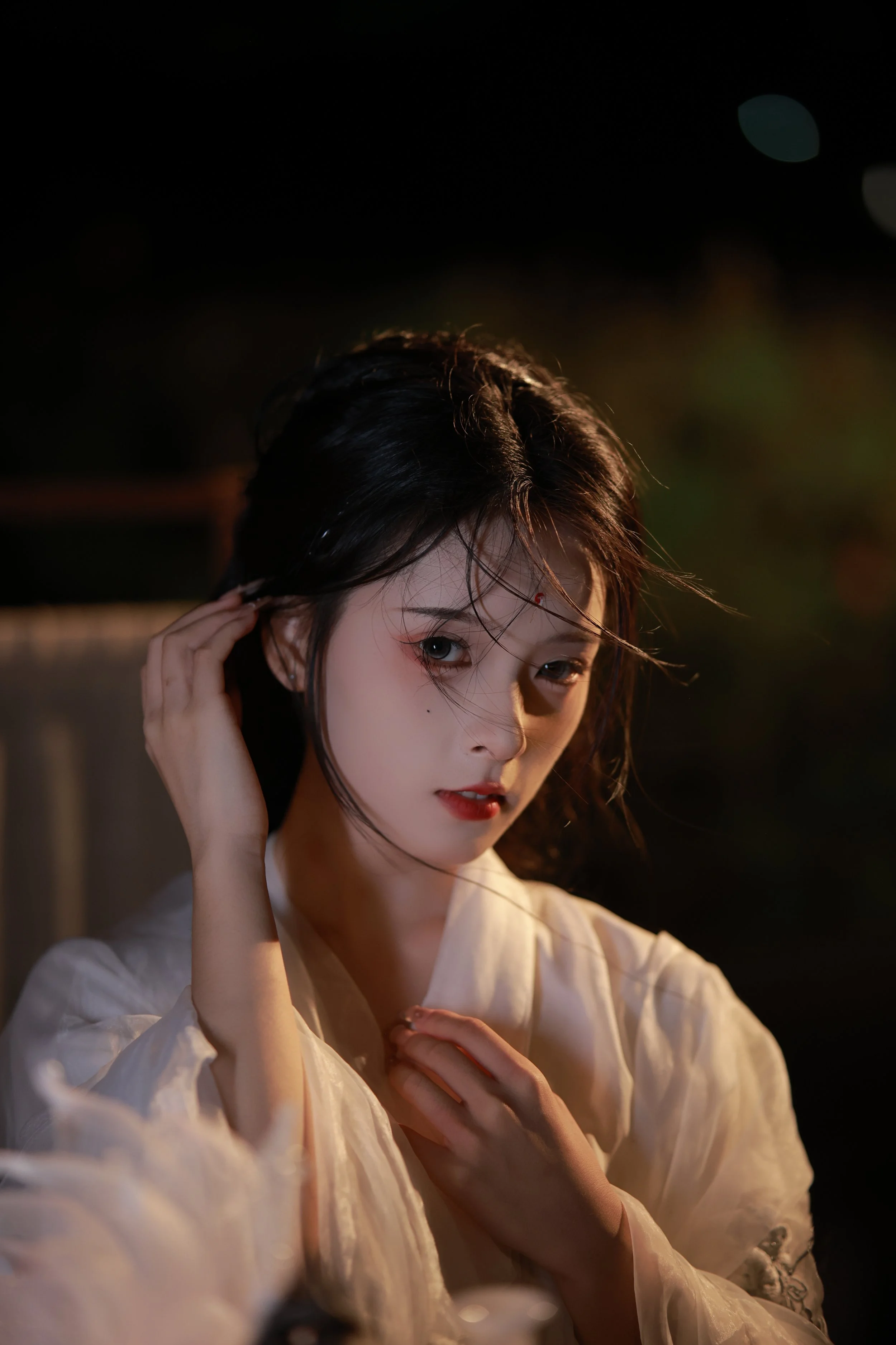 A woman with dark hair and red lipstick, wearing a light-colored satin robe, is gazing at the camera in a dimly lit environment.