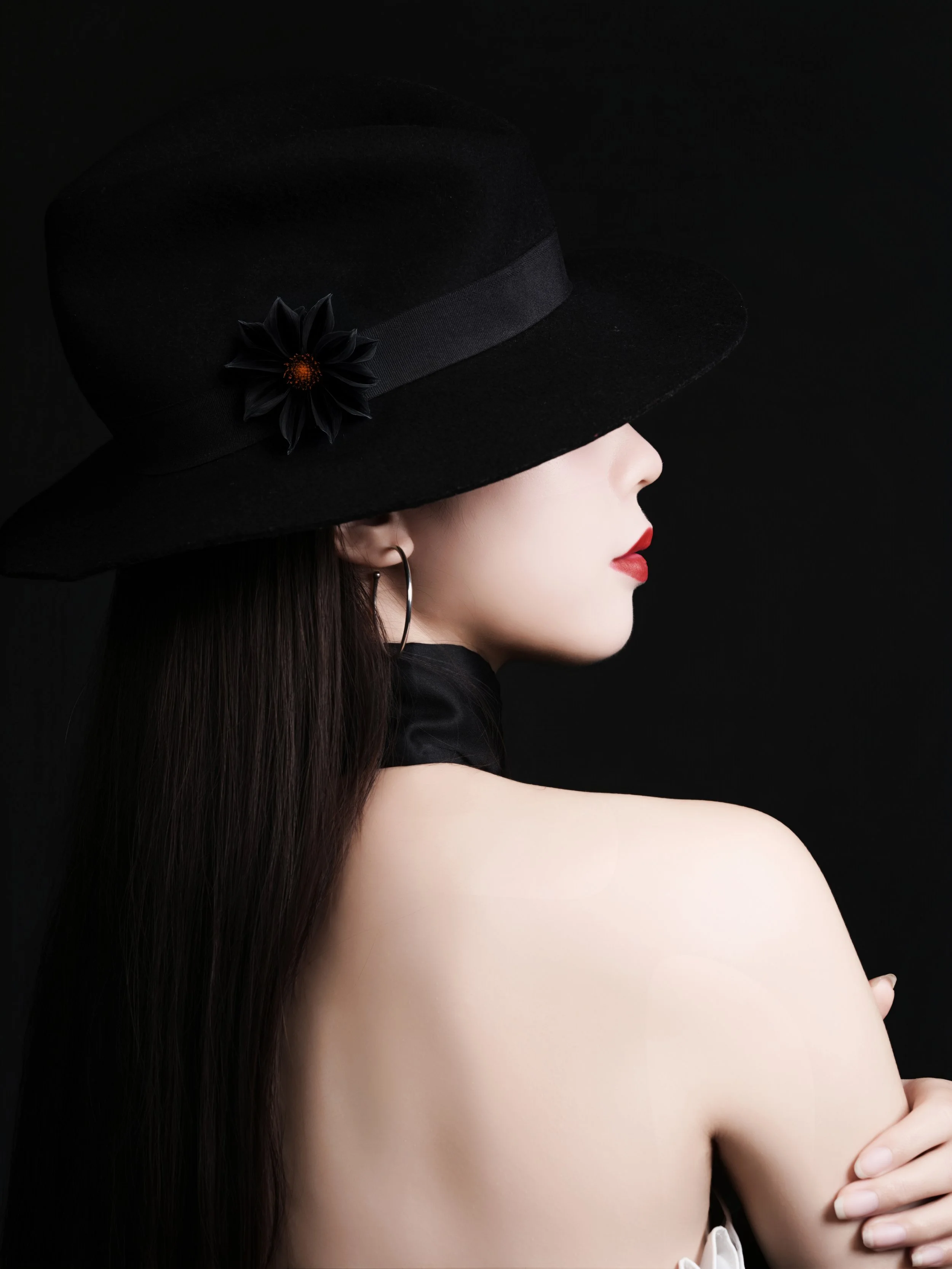 A woman with fair skin and dark hair, wearing a black wide-brimmed hat with a black flower, is shown in profile against a black background. She has red lipstick and hoop earrings, and is holding her shoulder with one hand.