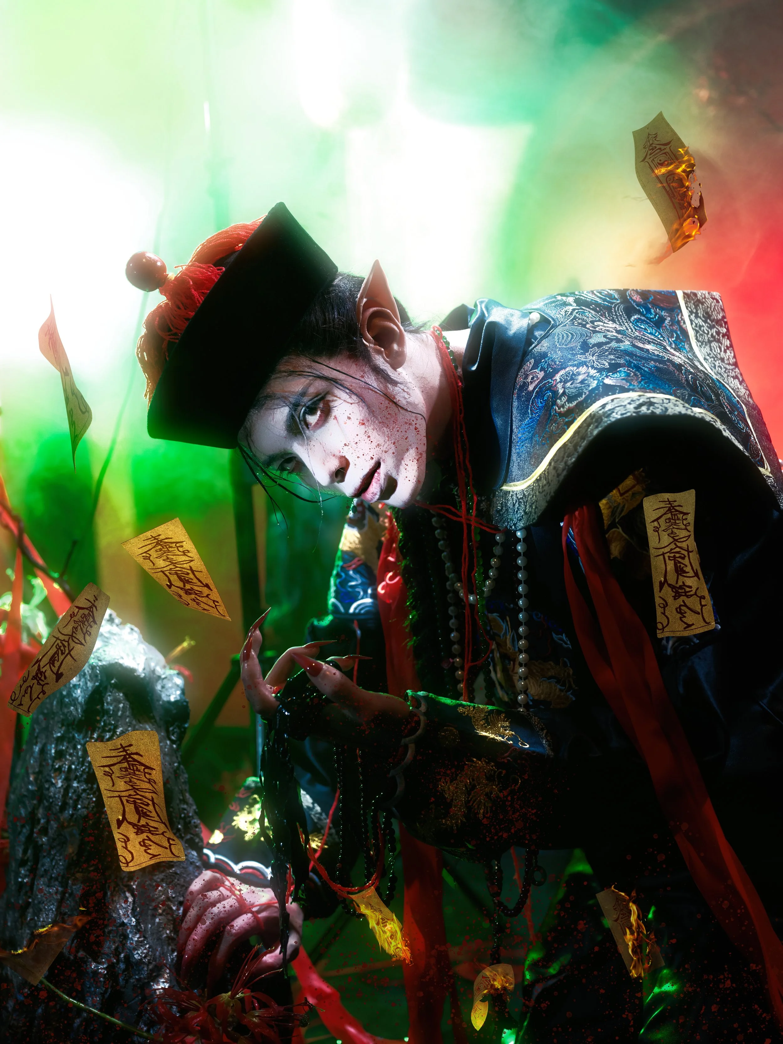 A person dressed in elaborate, dark-colored costume with intricate patterns, wearing a black hat with red tassels, and makeup resembling a zombie, surrounded by floating paper talismans and green and red lighting.