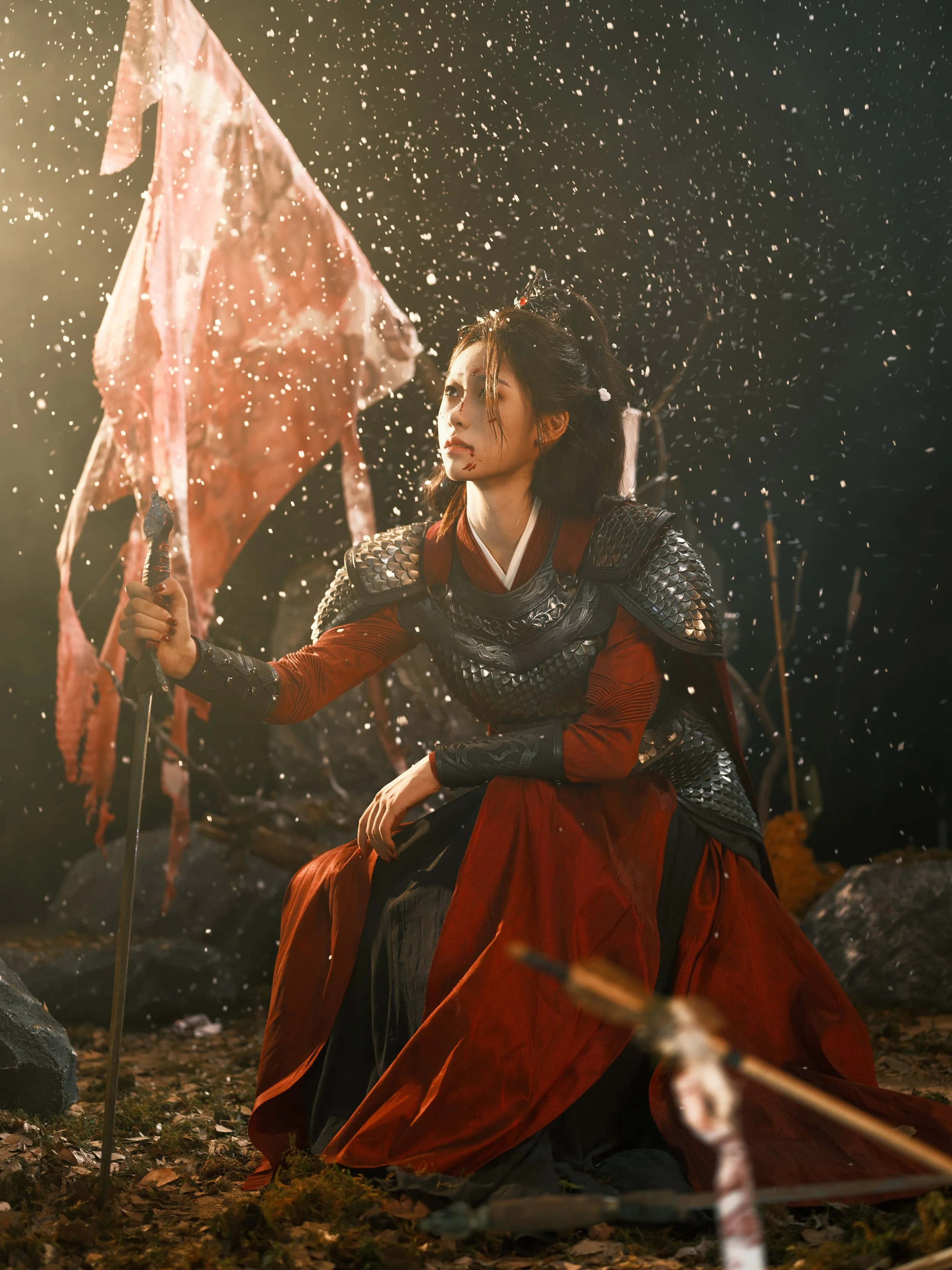 A woman in elaborate armor, with a flowing red dress, sitting on the ground in a dark outdoor setting, holding a flag, with debris and sparks around her.