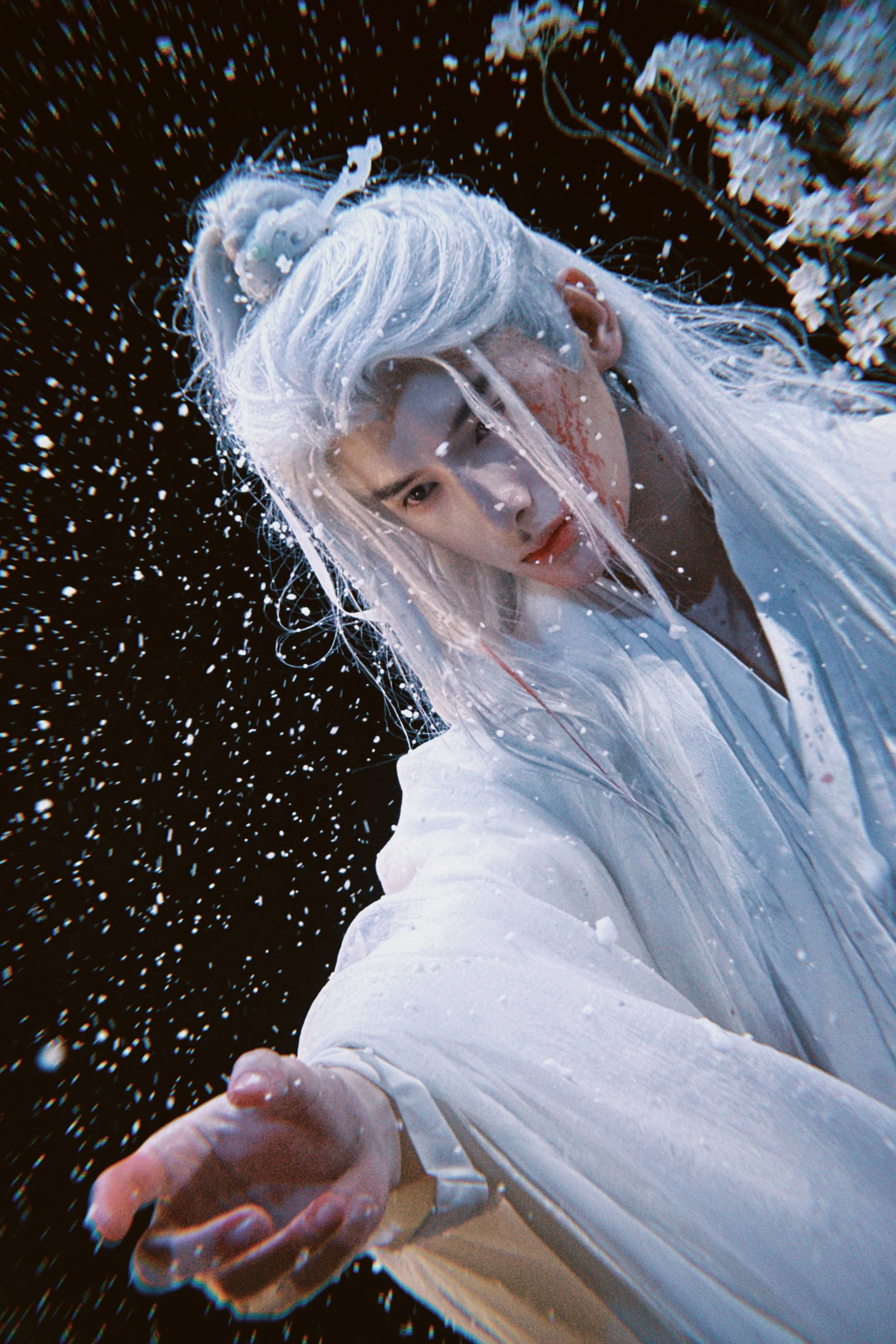 A person with long white hair shares a serious expression while surrounded by a shower of water droplets against a dark background, wearing traditional white clothing.