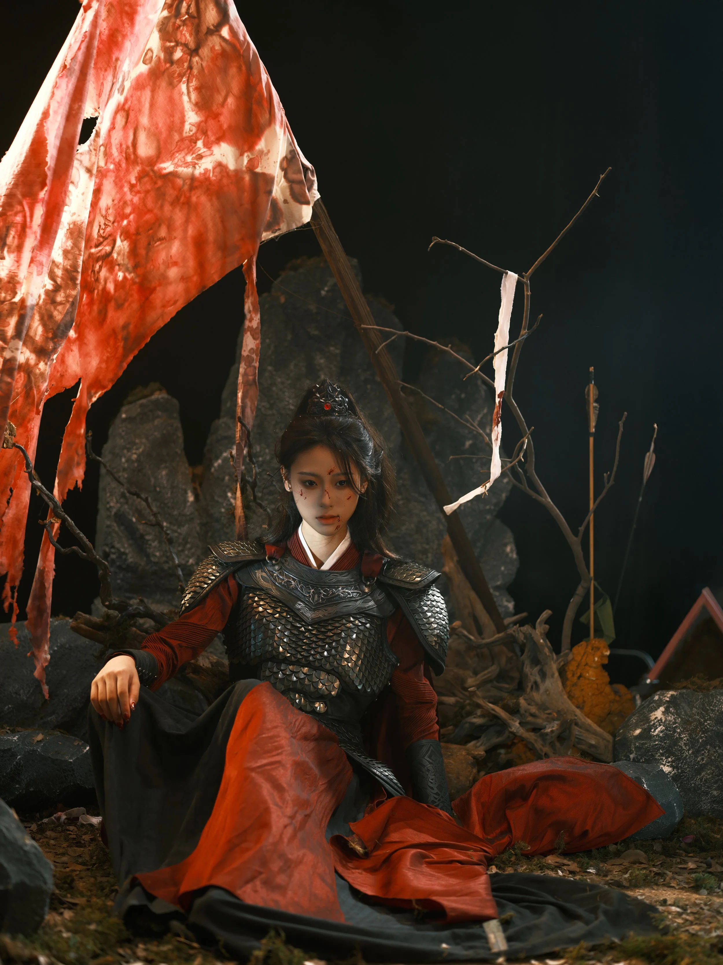 A woman dressed as a warrior in armor, sitting on the ground among rocks and branches with a dark background, a tattered red and white flag behind her.
