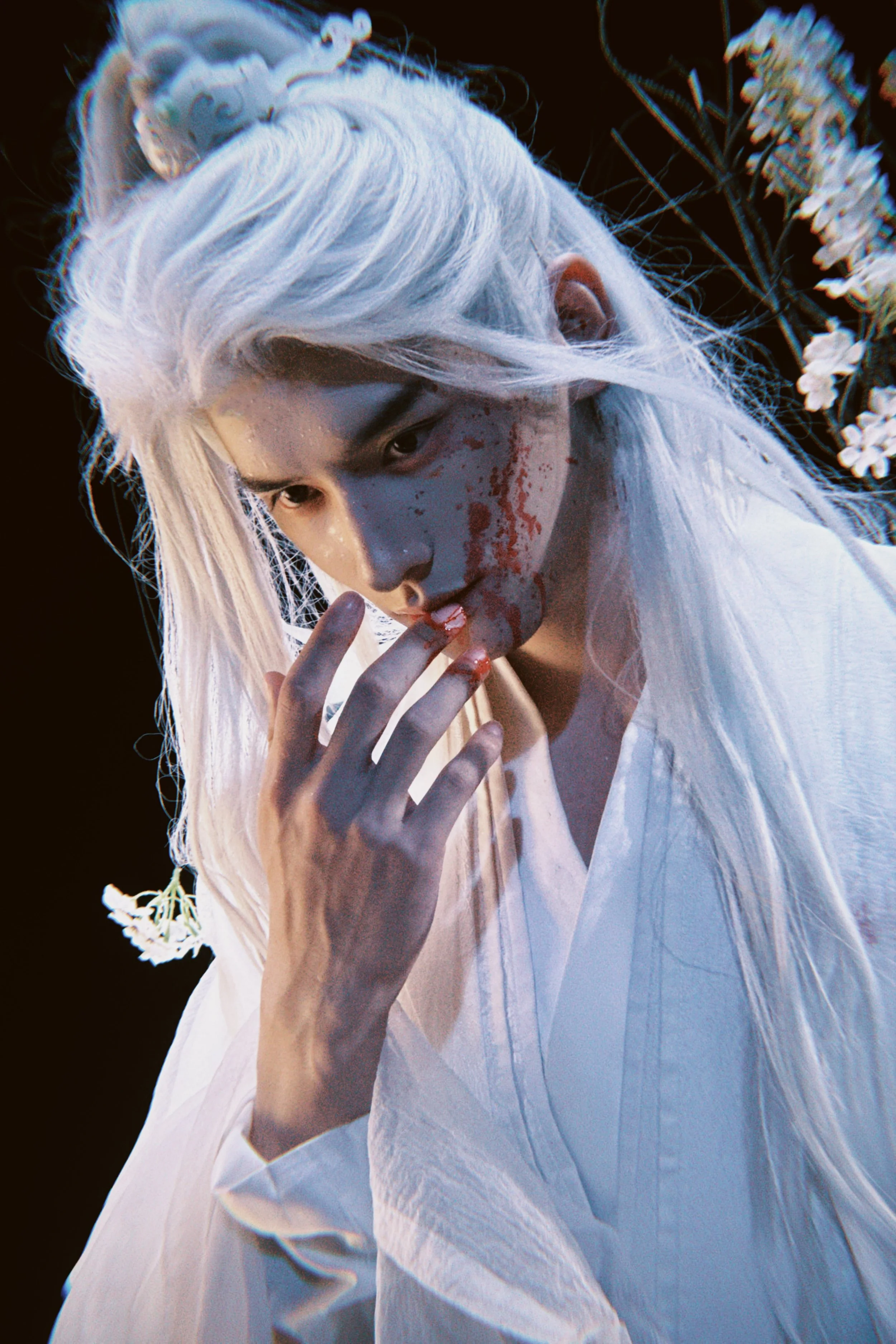 A person with long white hair, blood on their face and hand, and wearing a white traditional outfit, is gazing downward while touching their lips with fingers, with white flowers in the background.