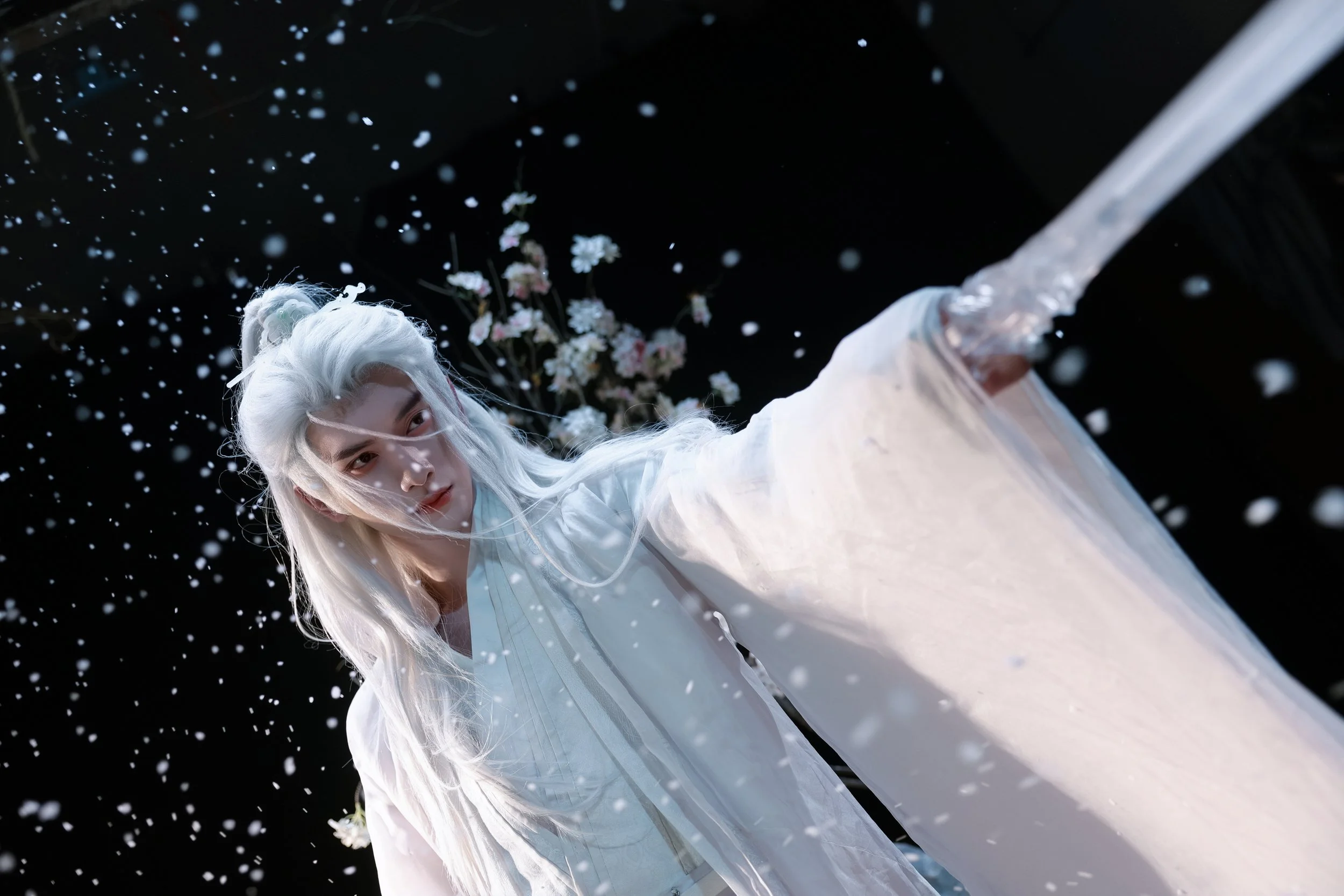 Person in traditional white costume with long white hair standing among falling flower petals with dark background and flowers in the distance.